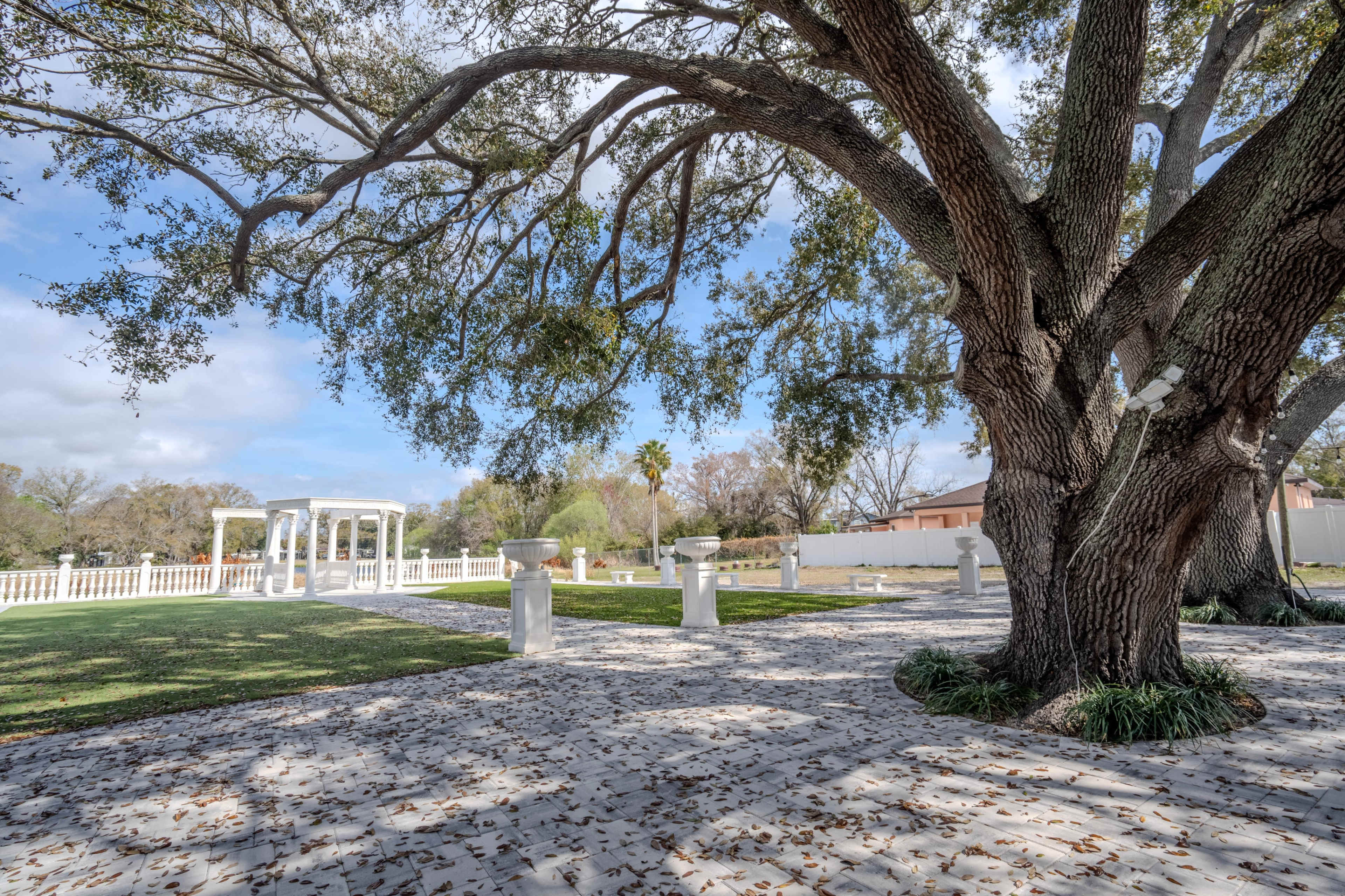 A large tree with spreading branches stands next to a grassy area that features a white gazebo and several columns on a paved surface.