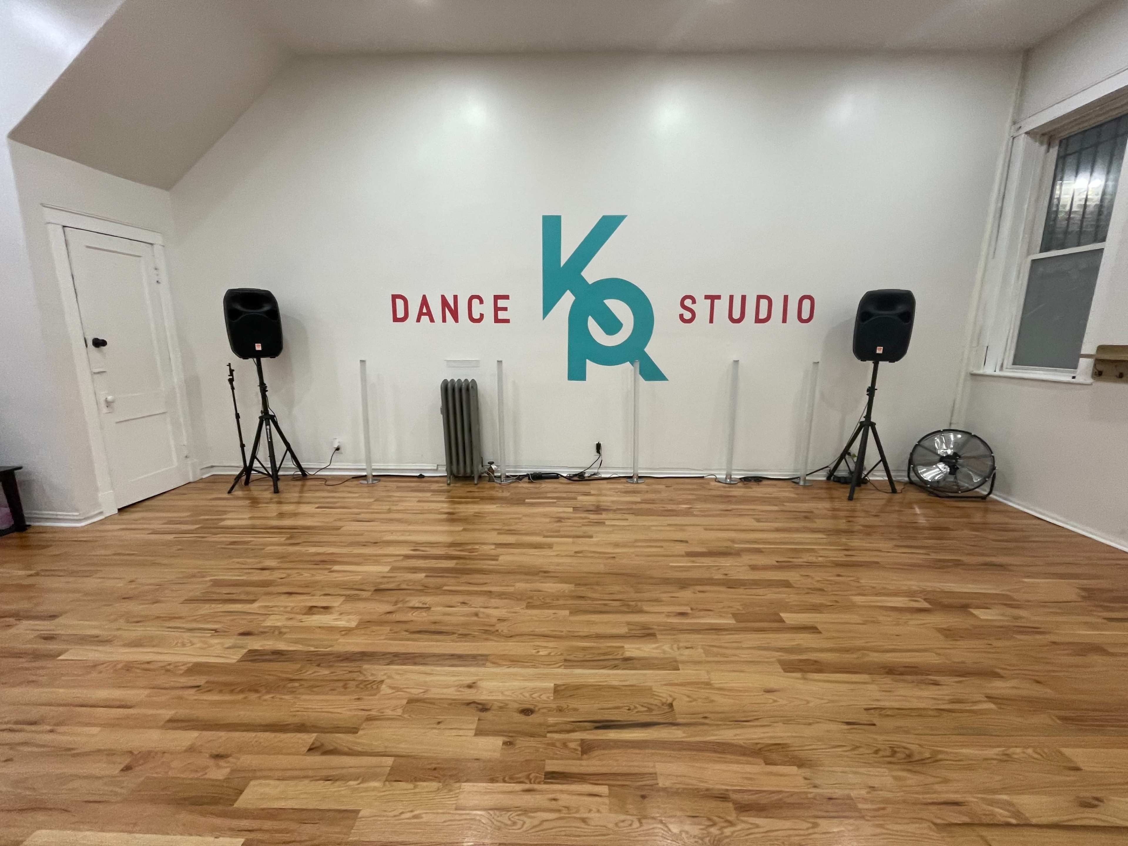 A spacious dance studio with wooden flooring, featuring large speakers on stands, a wall with the text "K Dance Studio," and a fan in the corner.