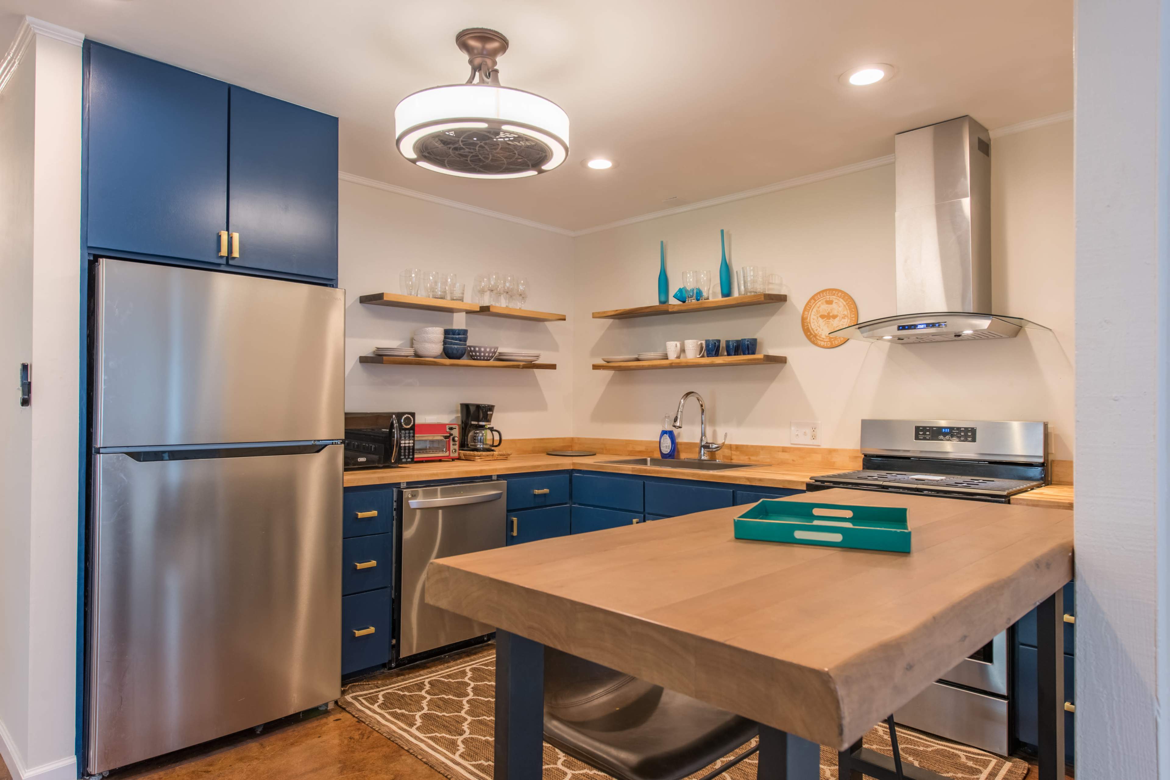 A kitchen with blue cabinets, stainless steel appliances, wooden shelves, and a central table.