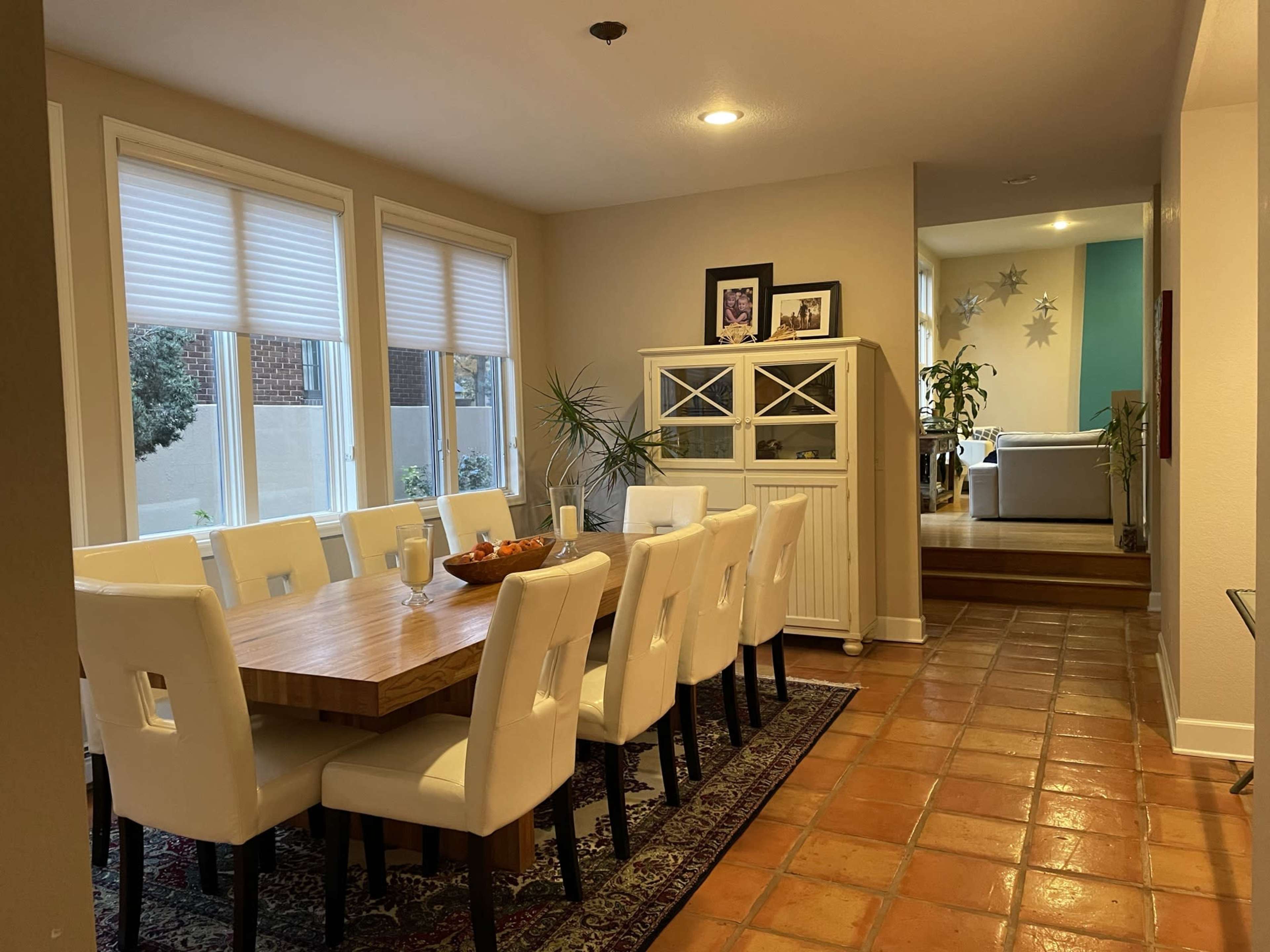 A spacious dining room features a large wooden table surrounded by white chairs, with a sideboard and windows offering views of a garden.