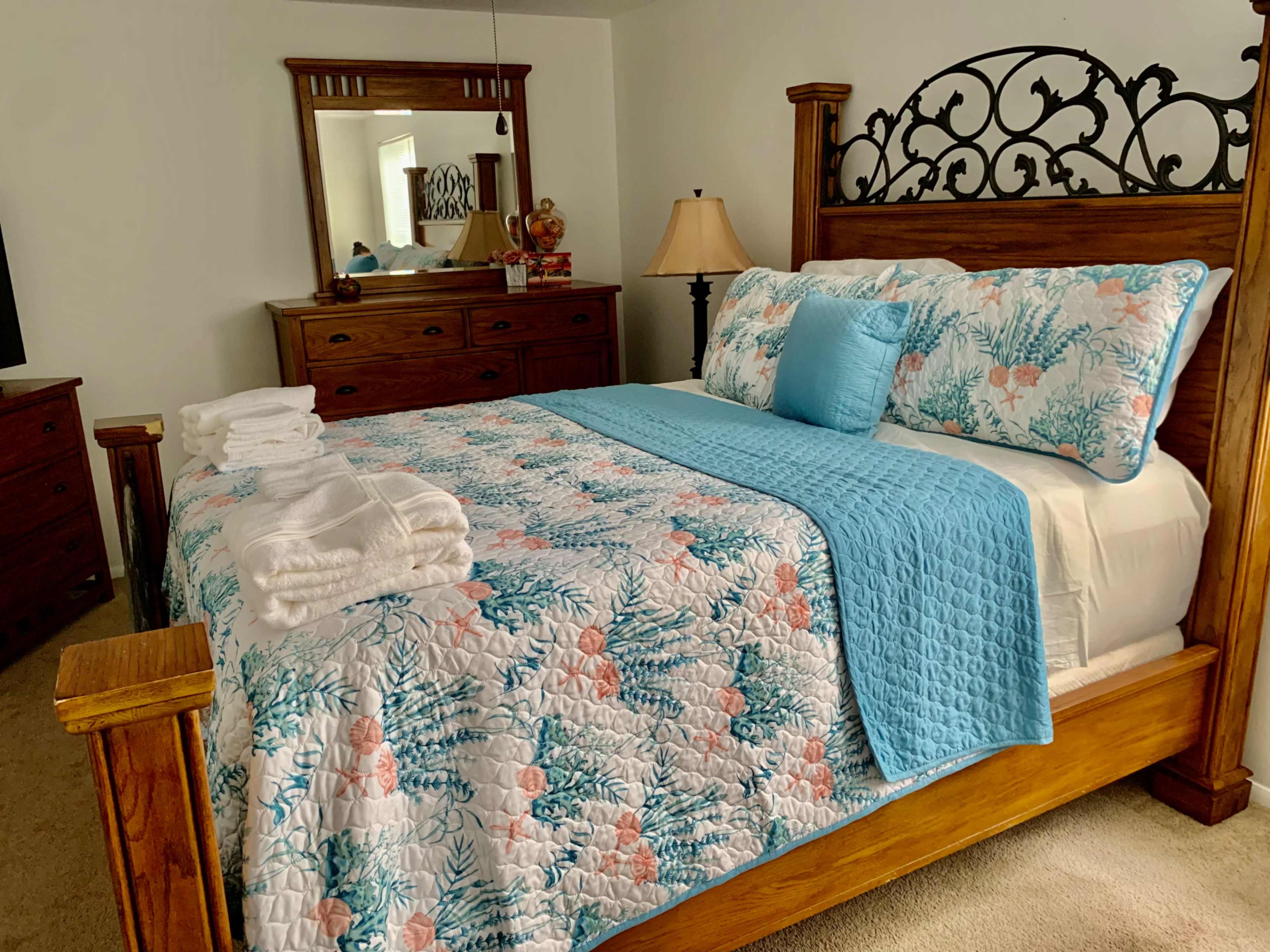 A neatly made bed with a floral-patterned comforter and a solid blue throw blanket is centered in a well-furnished bedroom featuring wooden furniture and a mirror.