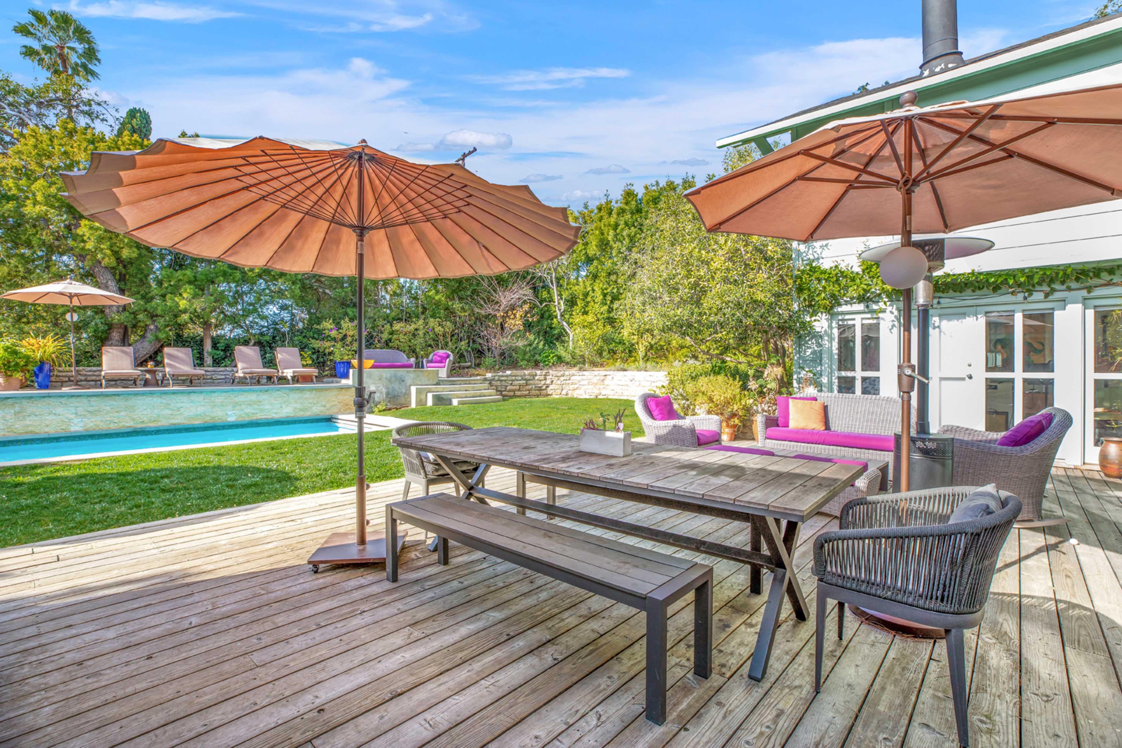 A wooden deck features a large table, black chairs, and two large umbrellas, overlooking a swimming pool surrounded by lounge chairs and greenery.