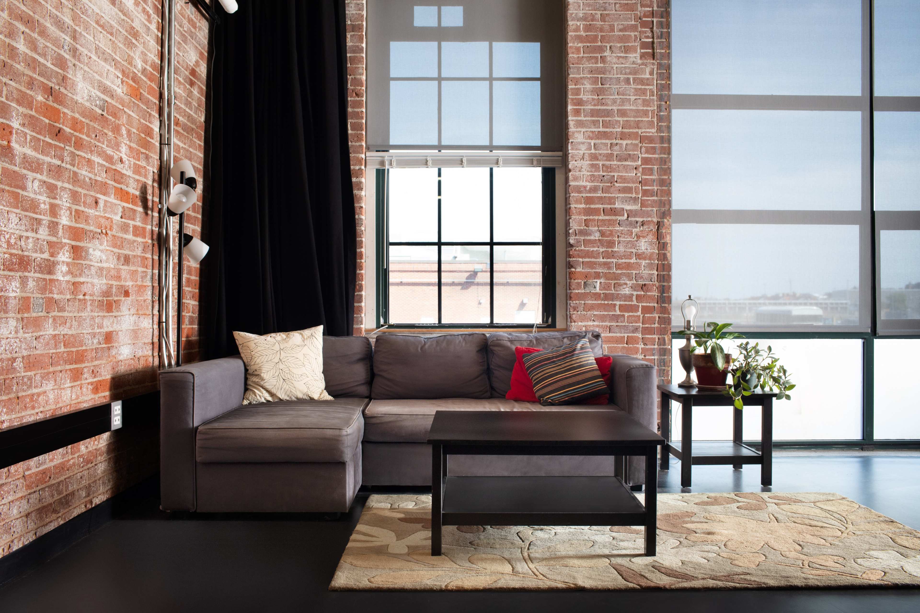 A modern living room featuring a gray sectional sofa, a coffee table, and a large window with sheer curtains against a backdrop of exposed brick walls.