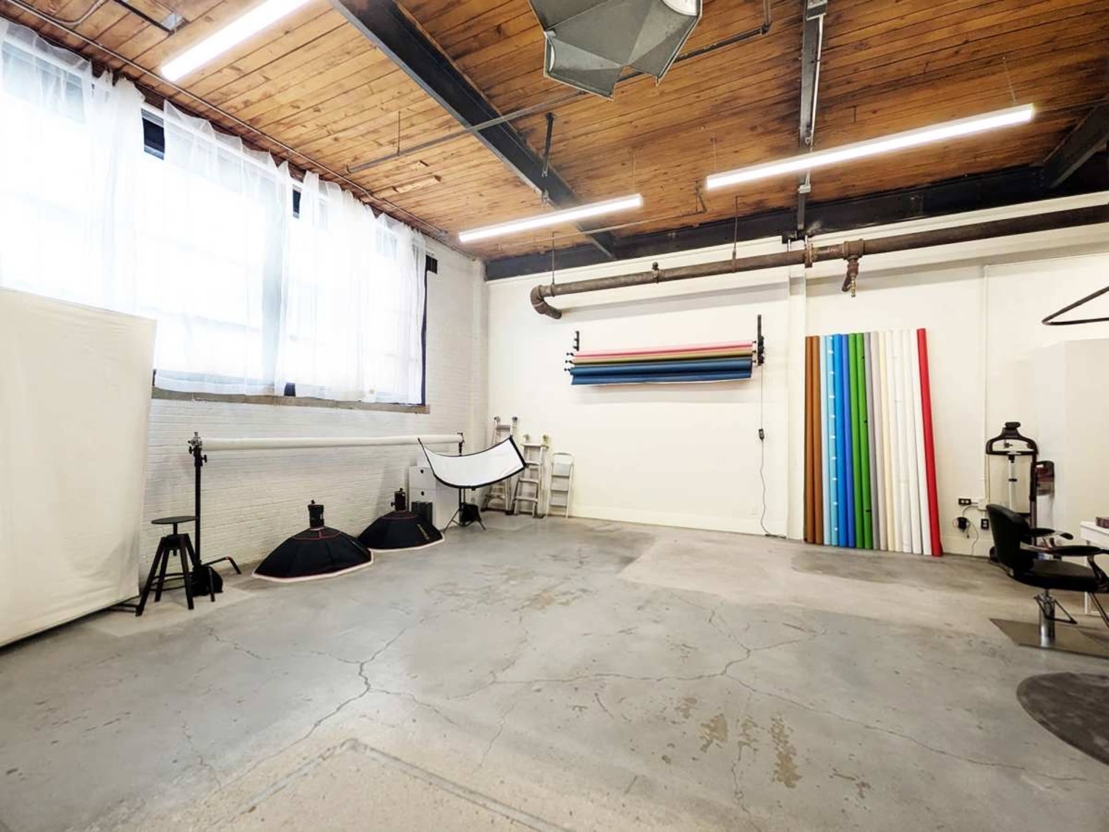 The image depicts a spacious photography studio featuring muted concrete flooring, large windows with sheer curtains, and various equipment arranged against the walls, including backdrop rolls and lighting stands.