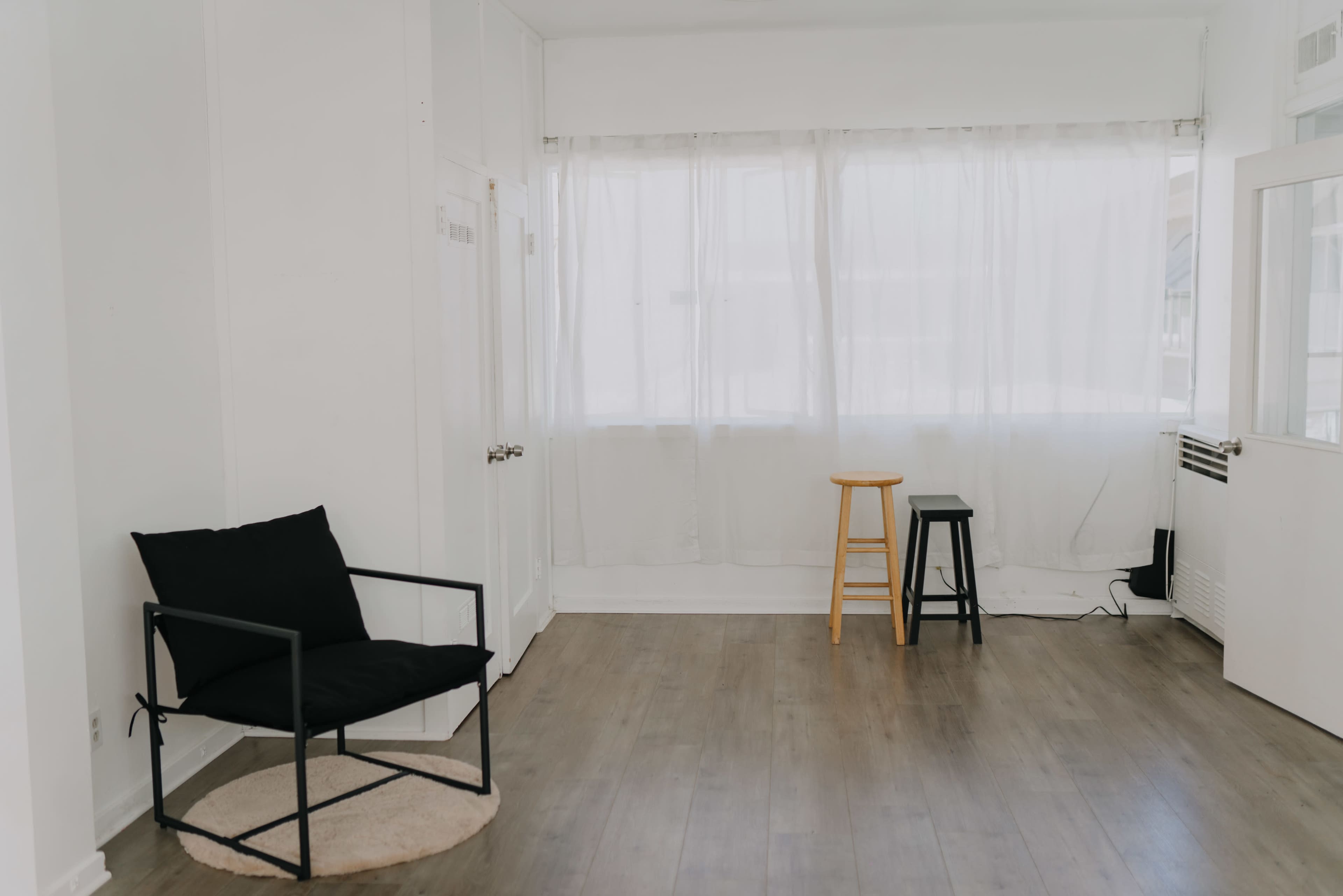 The image shows a bright, minimally furnished room with a black chair, a wooden stool, and a black stool, all positioned on a light hardwood floor.