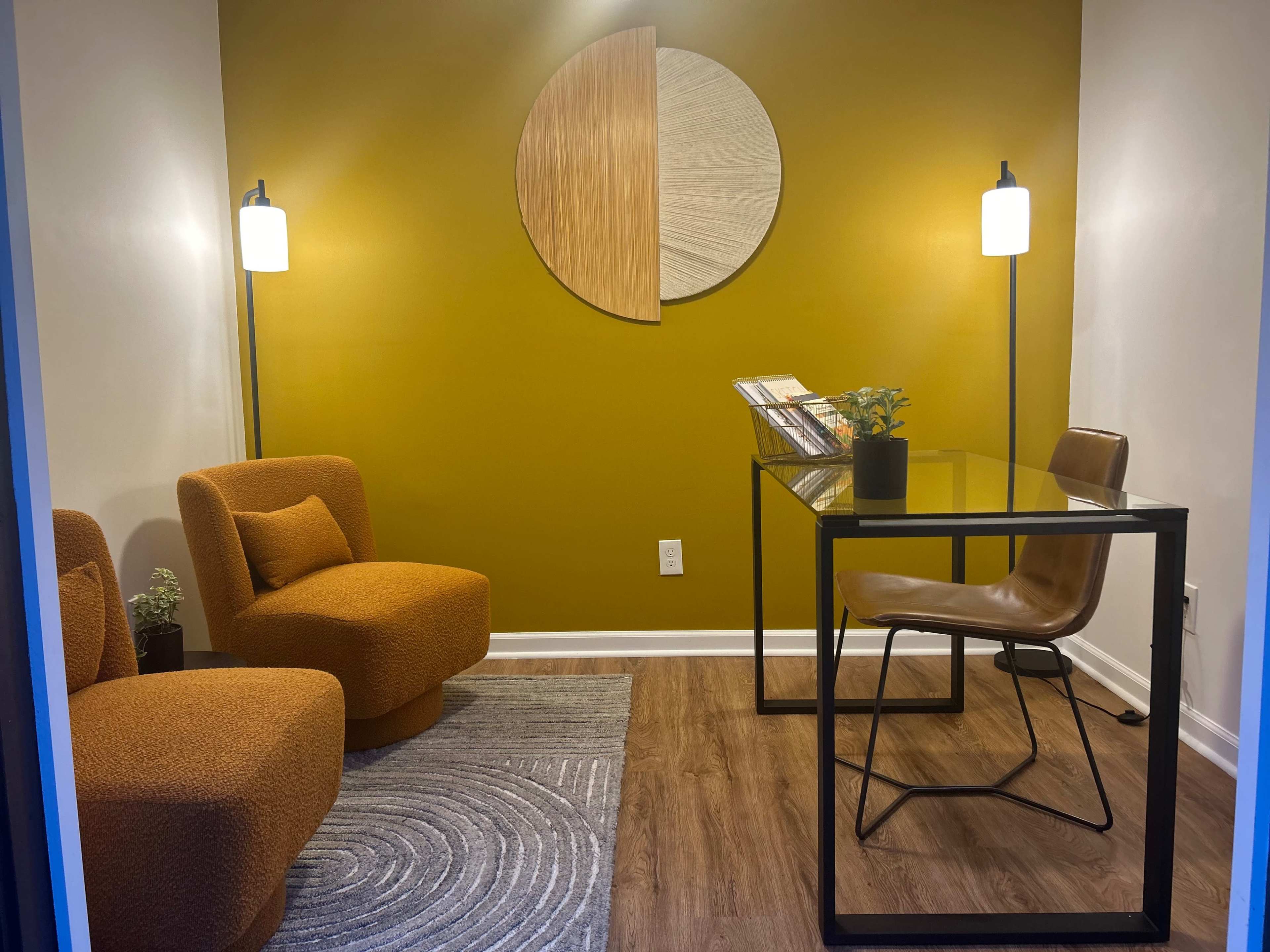 A small modern office space with two orange upholstered chairs, a glass desk with a brown chair, and a decorative wall featuring a circular artwork on a mustard-colored background.