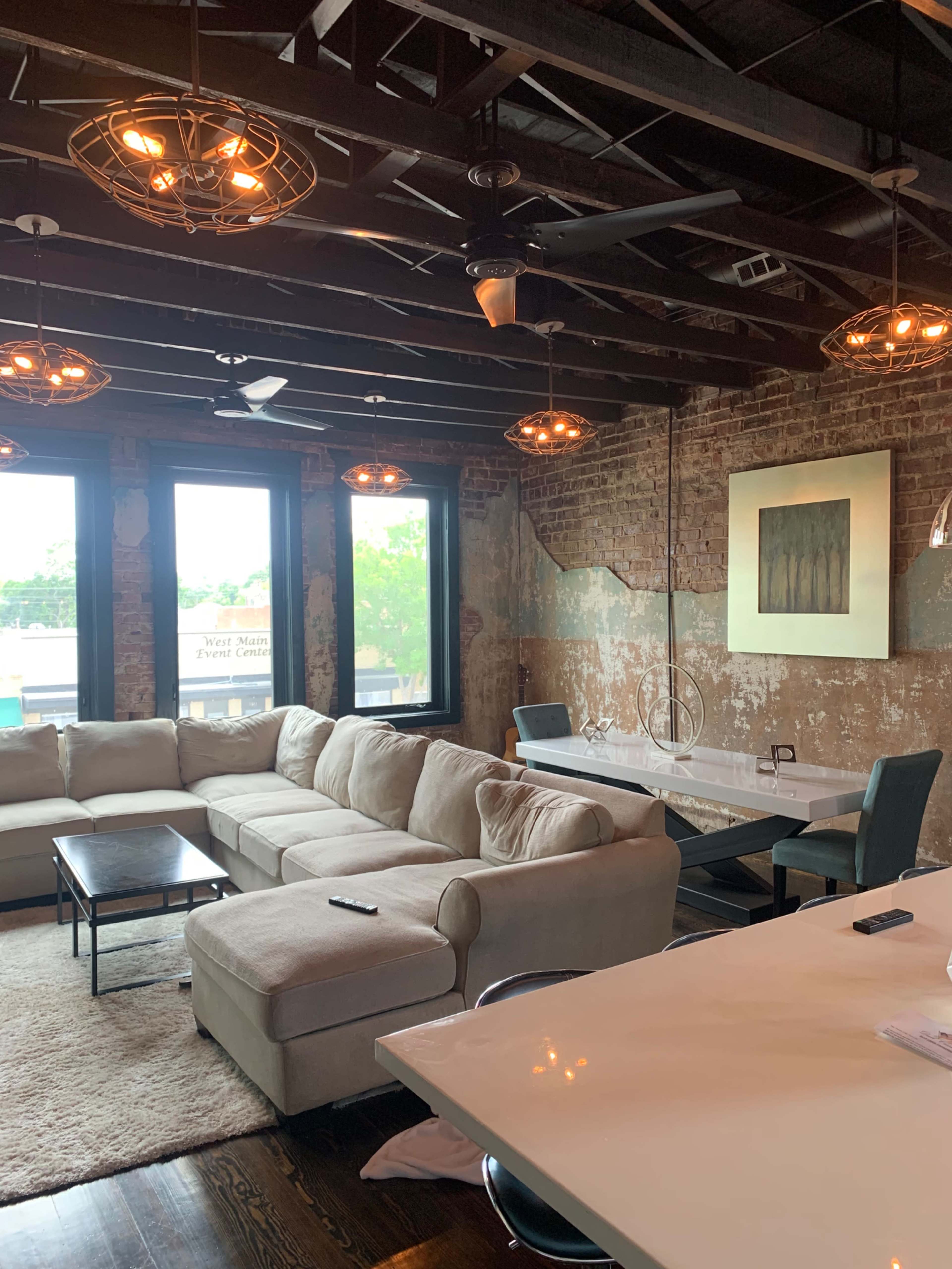 The image shows a spacious living room with exposed brick walls, large windows, a light-colored sectional sofa, and a dining table.