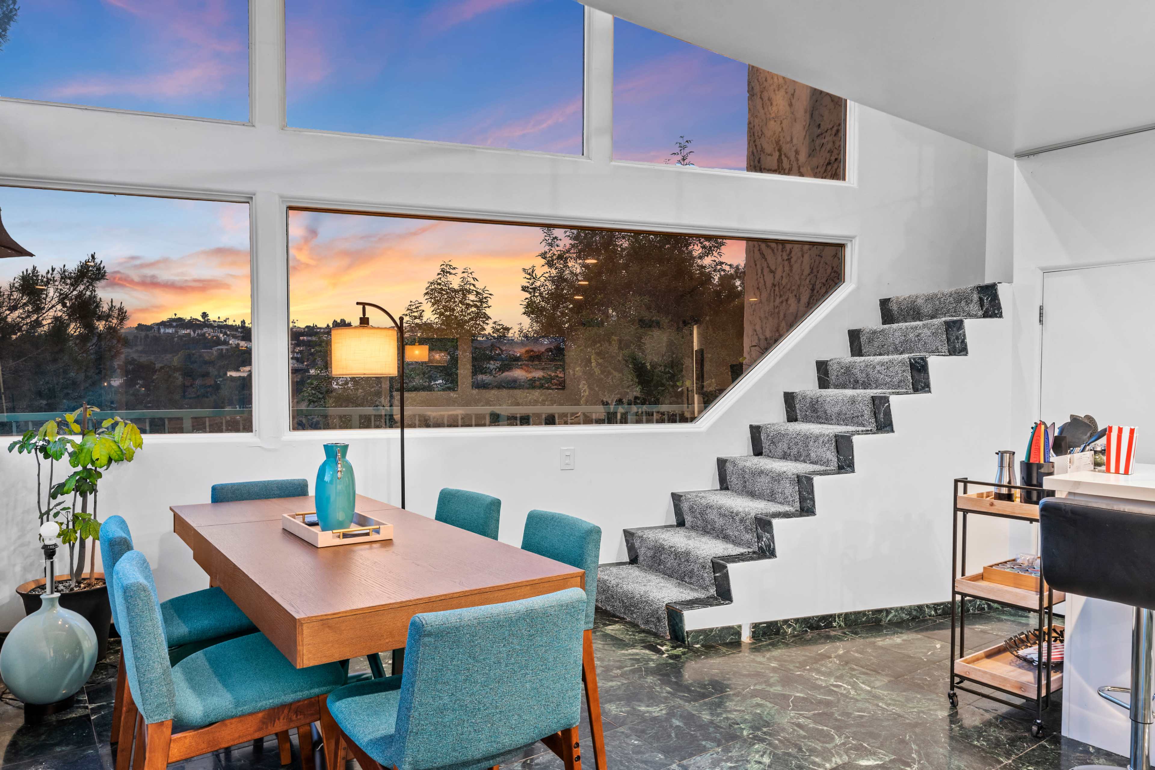 A dining area with a wooden table and teal chairs is adjacent to large windows displaying a sunset, while a staircase with a gray carpet leads to an upper level.
