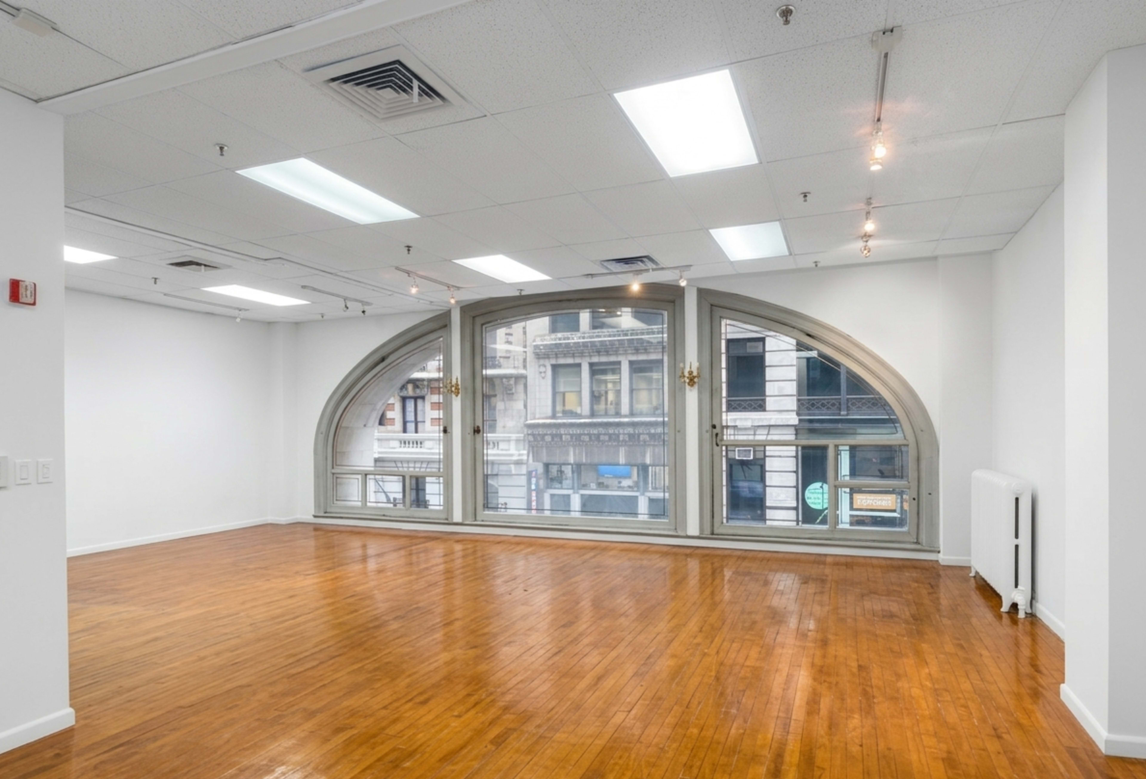 The image depicts a spacious, empty room with wooden floors and large arched windows that provide street-level views of an adjacent building.