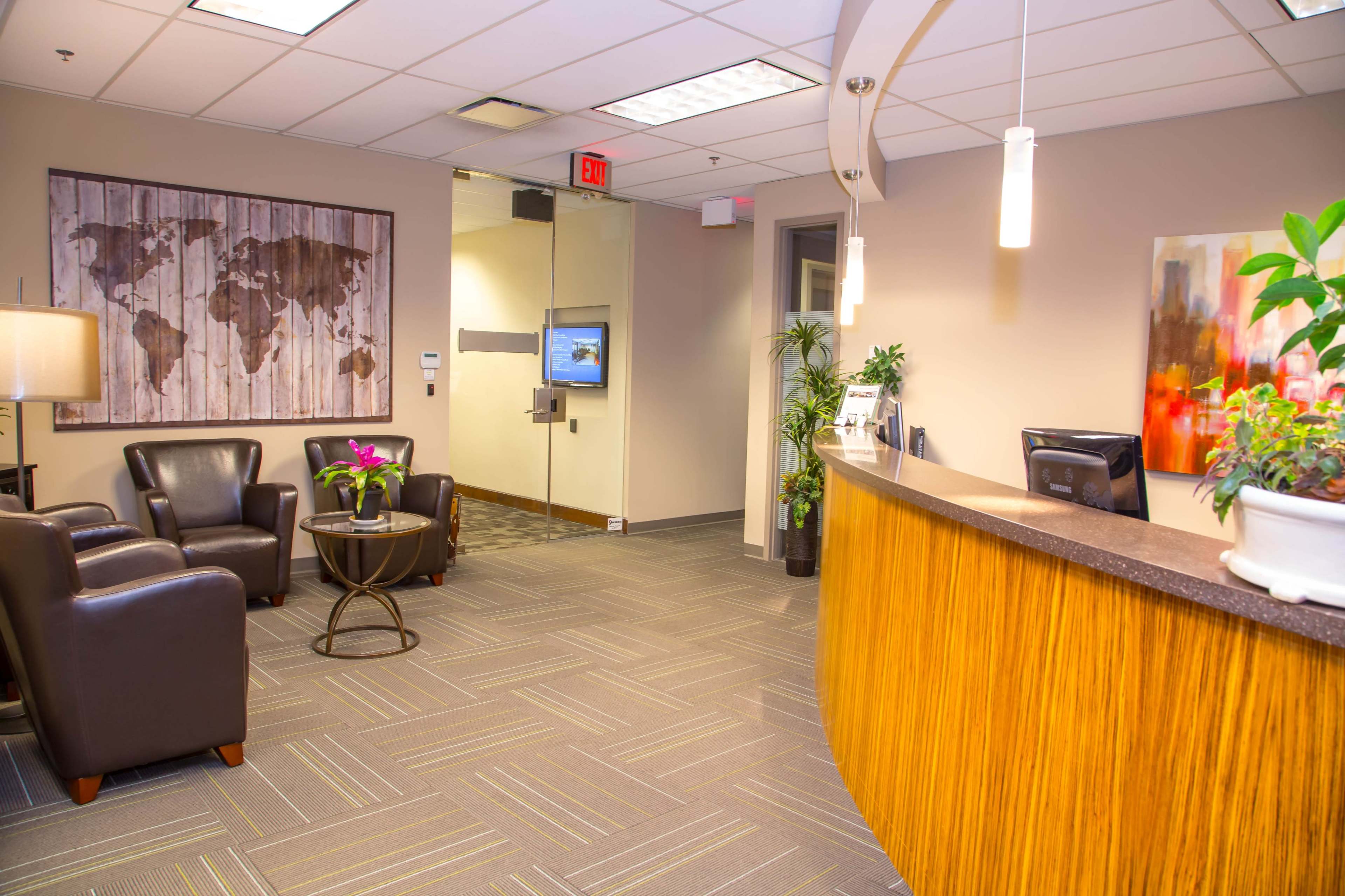 A modern reception area with a curved wooden desk, leather chairs, a coffee table, a mural of a world map on the wall, and plants in pots.