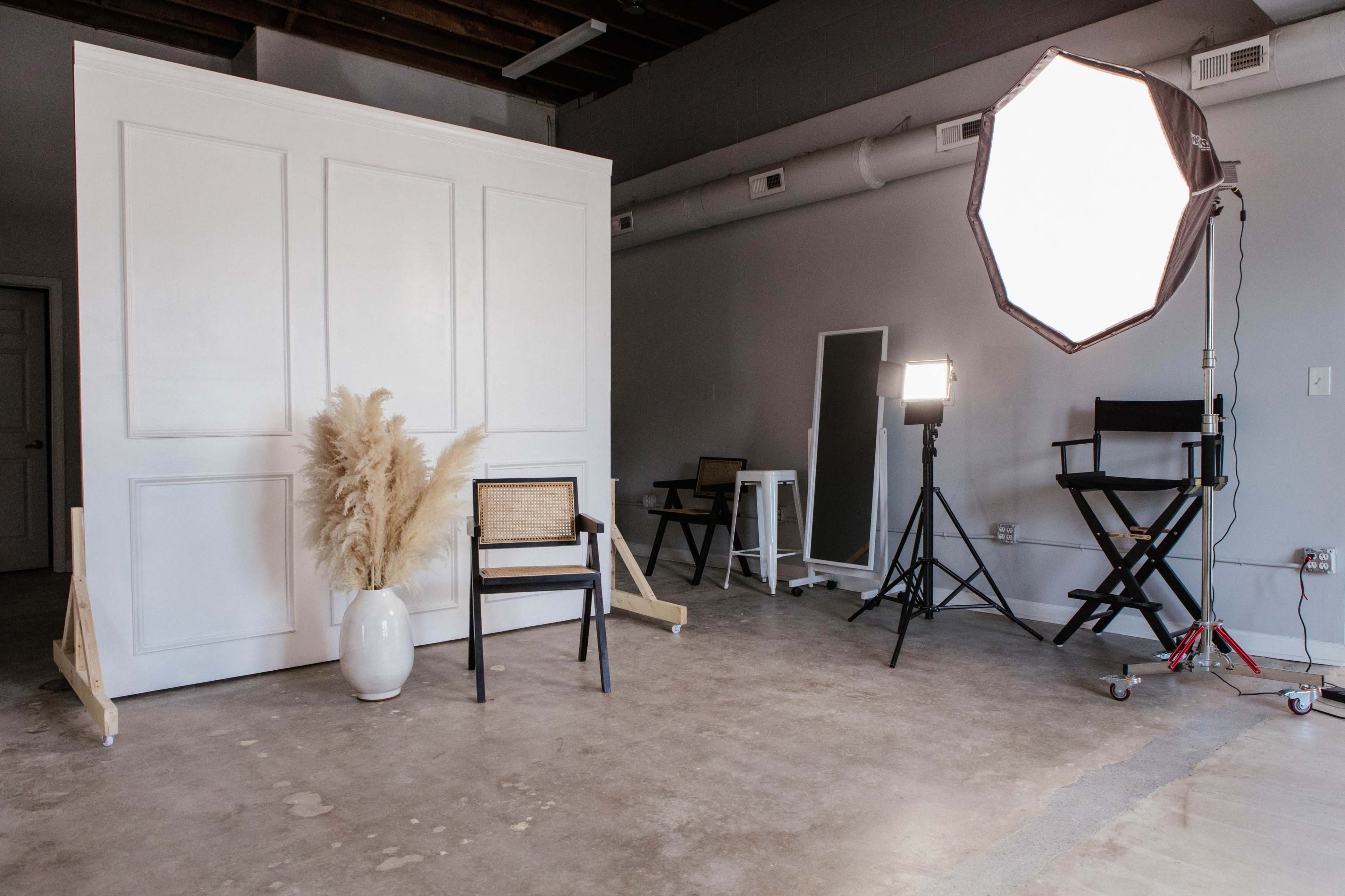 The image shows a minimalist photo studio setup with a white backdrop, a chair, and lighting equipment positioned around a bare concrete floor.