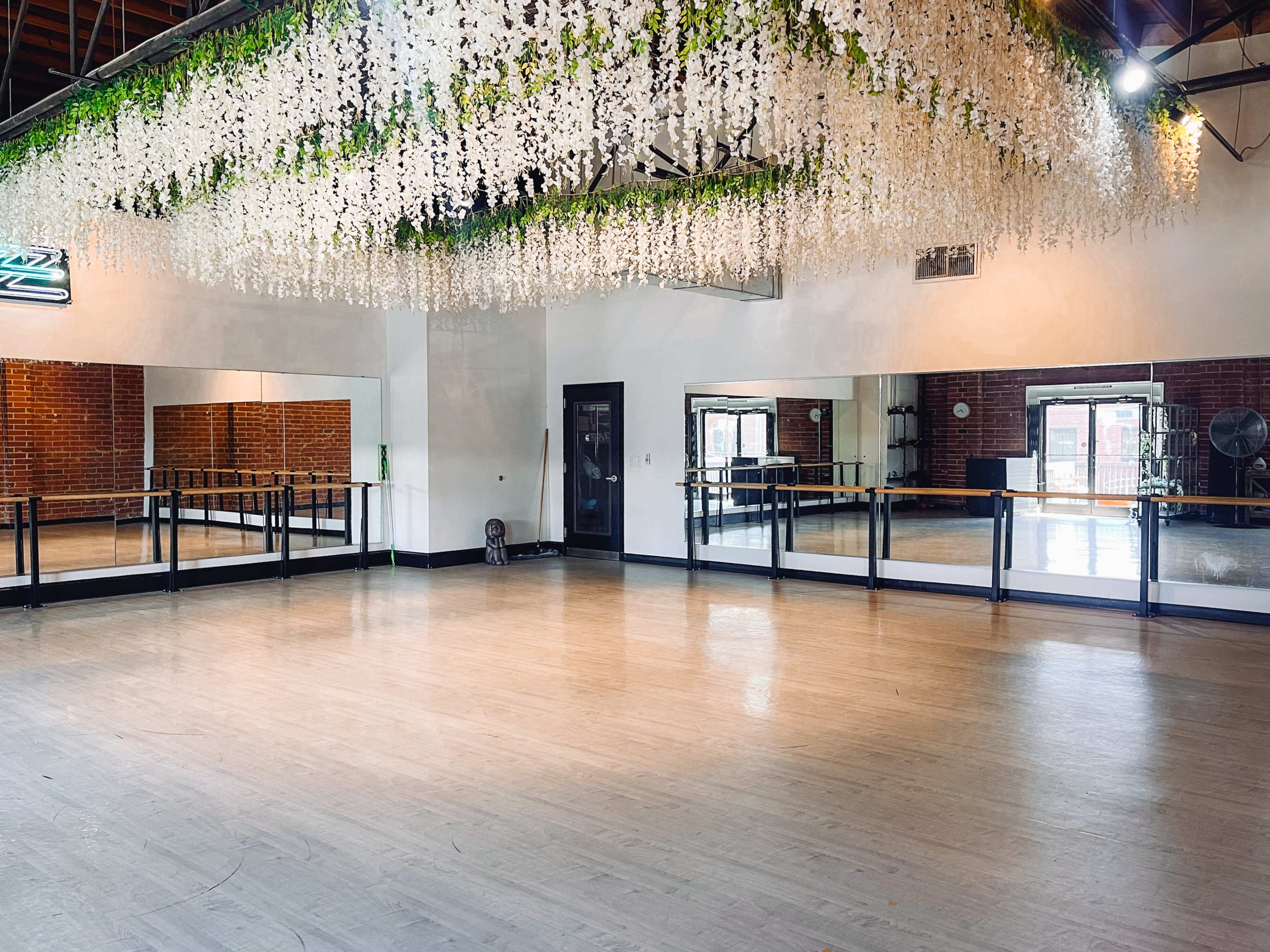 The image shows a spacious dance studio with a polished wooden floor, large mirrors on one wall, and a decorated ceiling featuring hanging floral arrangements.