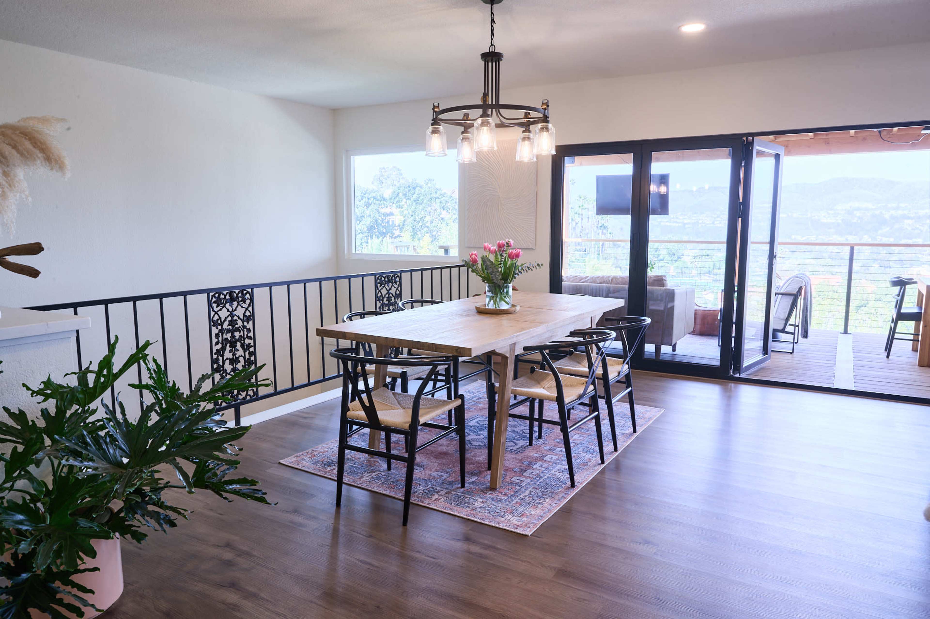 A dining area features a wooden table surrounded by black chairs, with a flower arrangement at its center, and large windows overlooking an outdoor space.