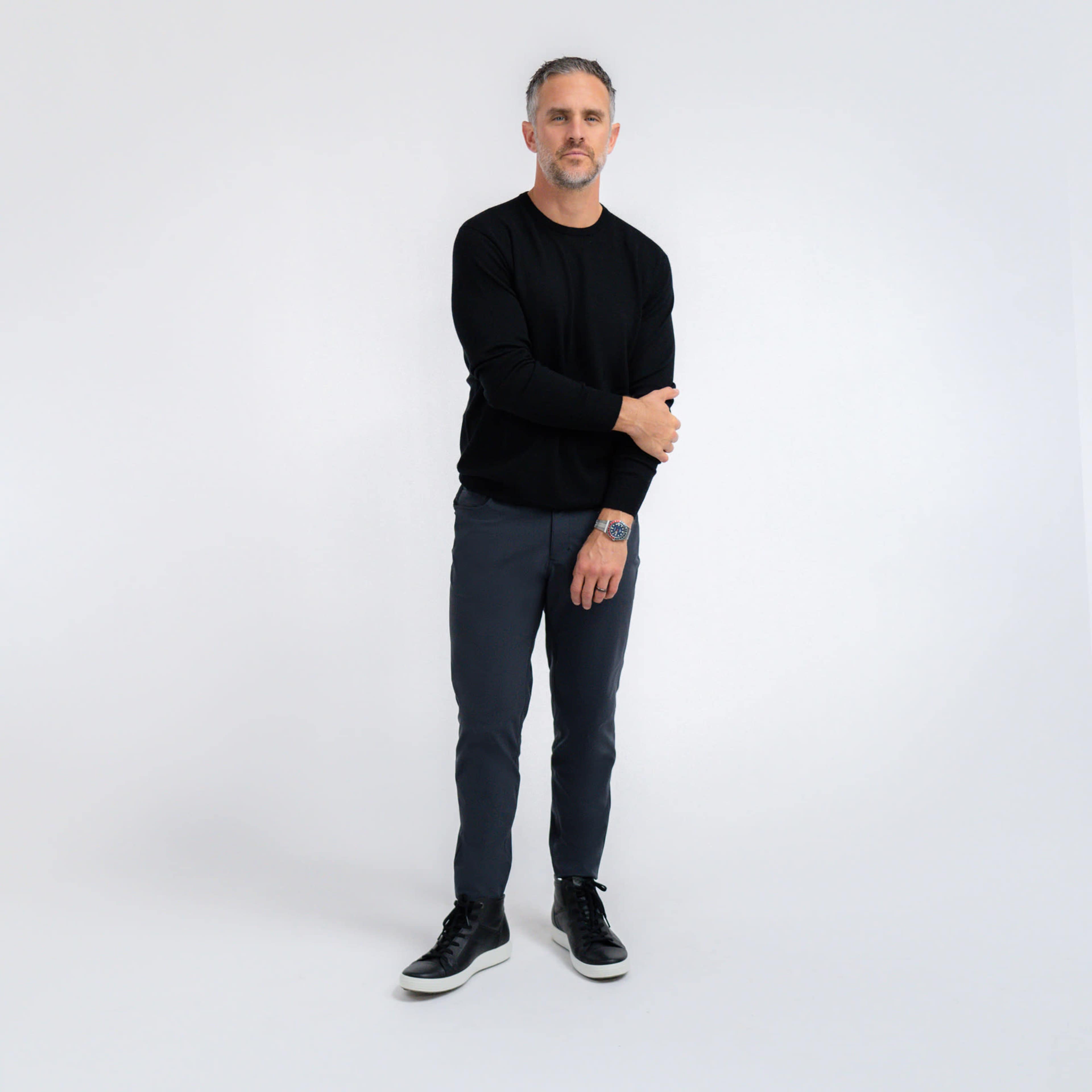A man stands facing the camera, wearing a black sweater, gray pants, and black sneakers.