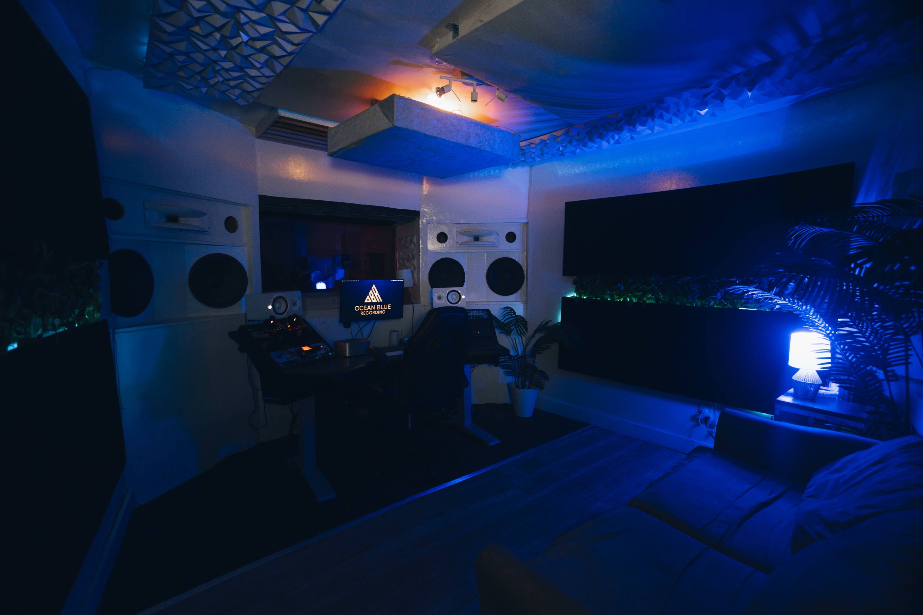 The image shows a dimly lit music studio featuring large speakers, a computer setup with a monitor, a couch, and ambient blue lighting.