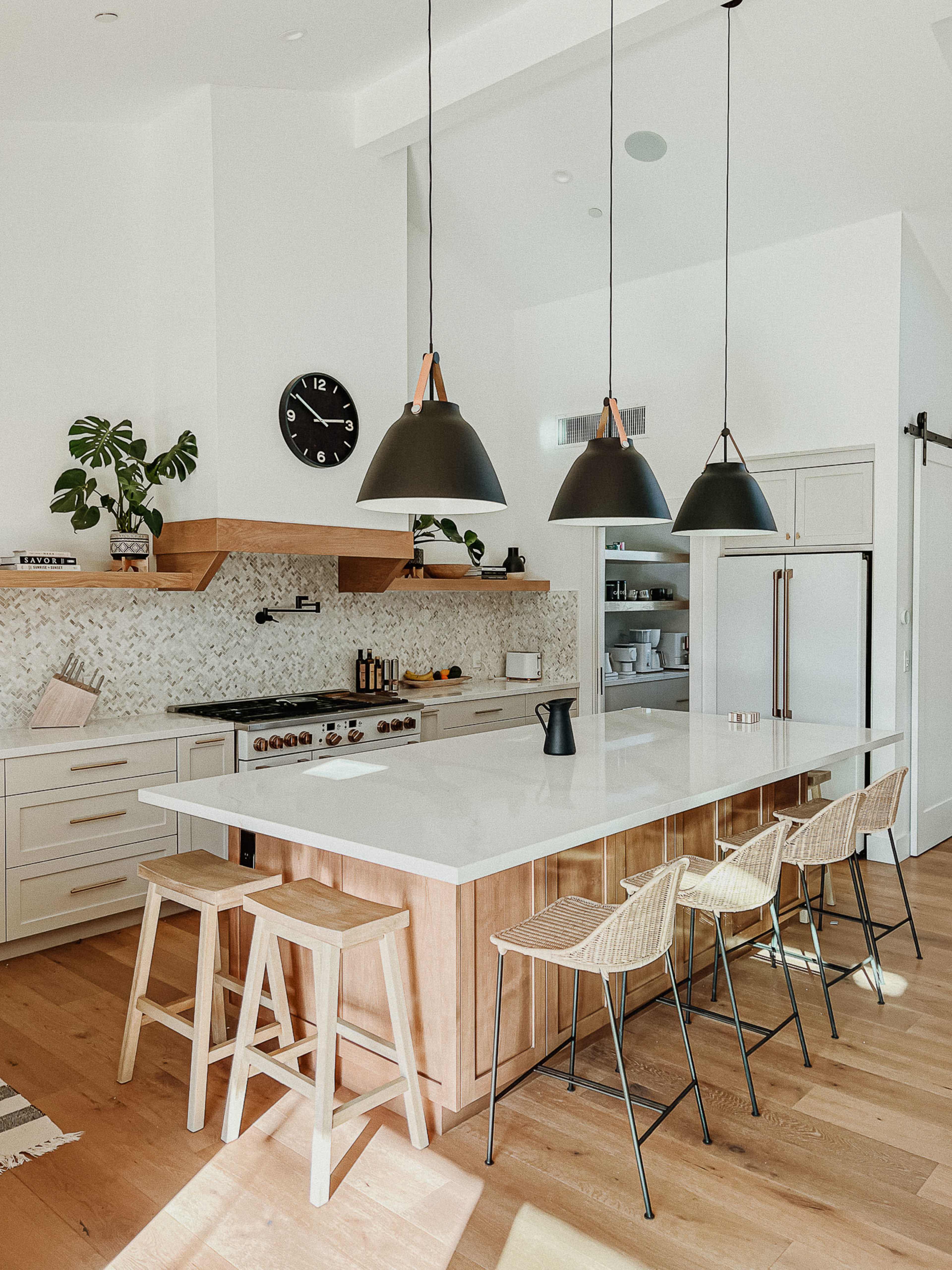 The kitchen features a large island with three black pendant lights, surrounded by modern bar stools, and is equipped with a stove, a decorative backsplash, and open shelving.