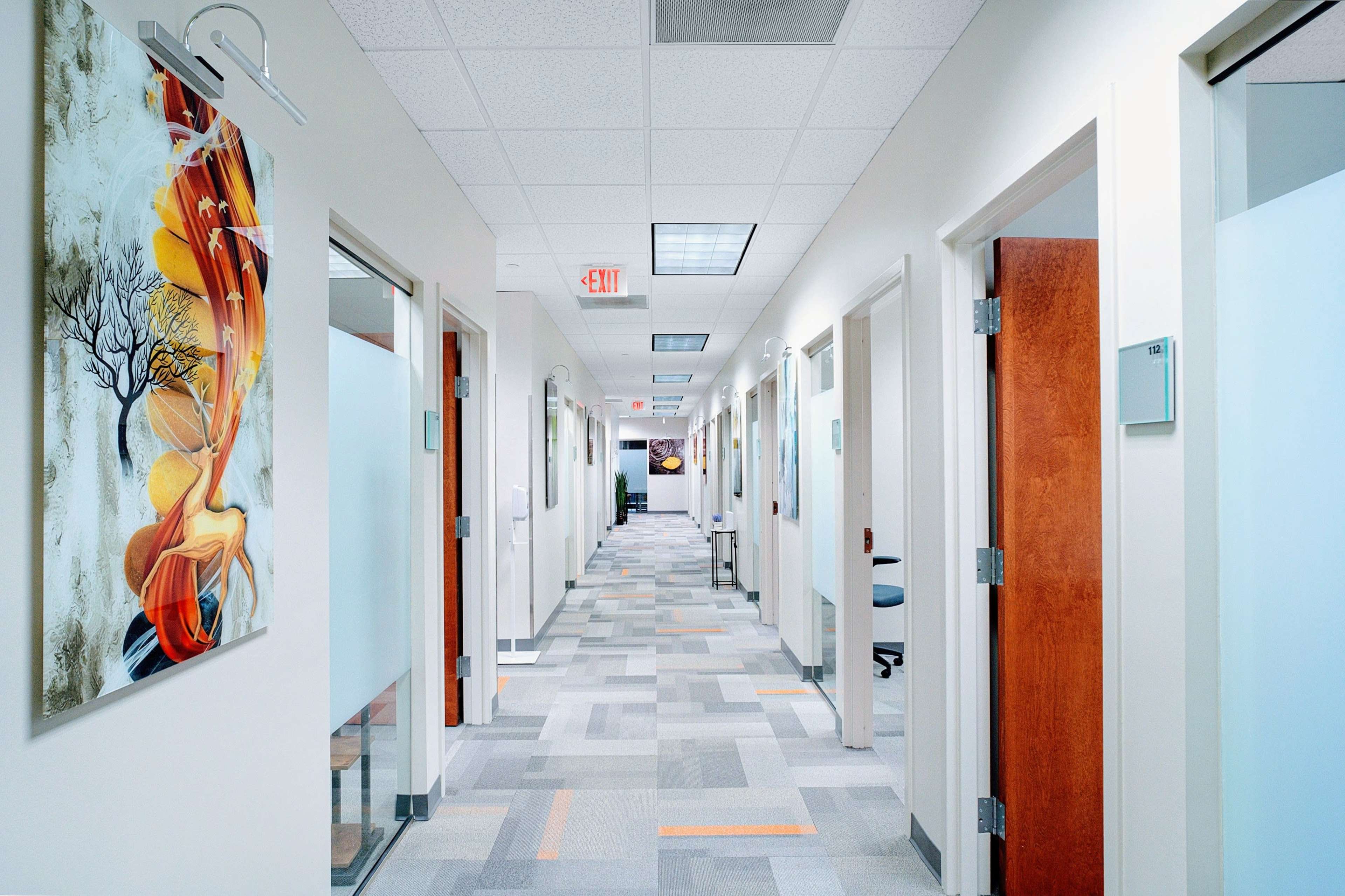 A well-lit hallway features multiple closed doors on either side, with a colorful piece of art hanging on one wall.