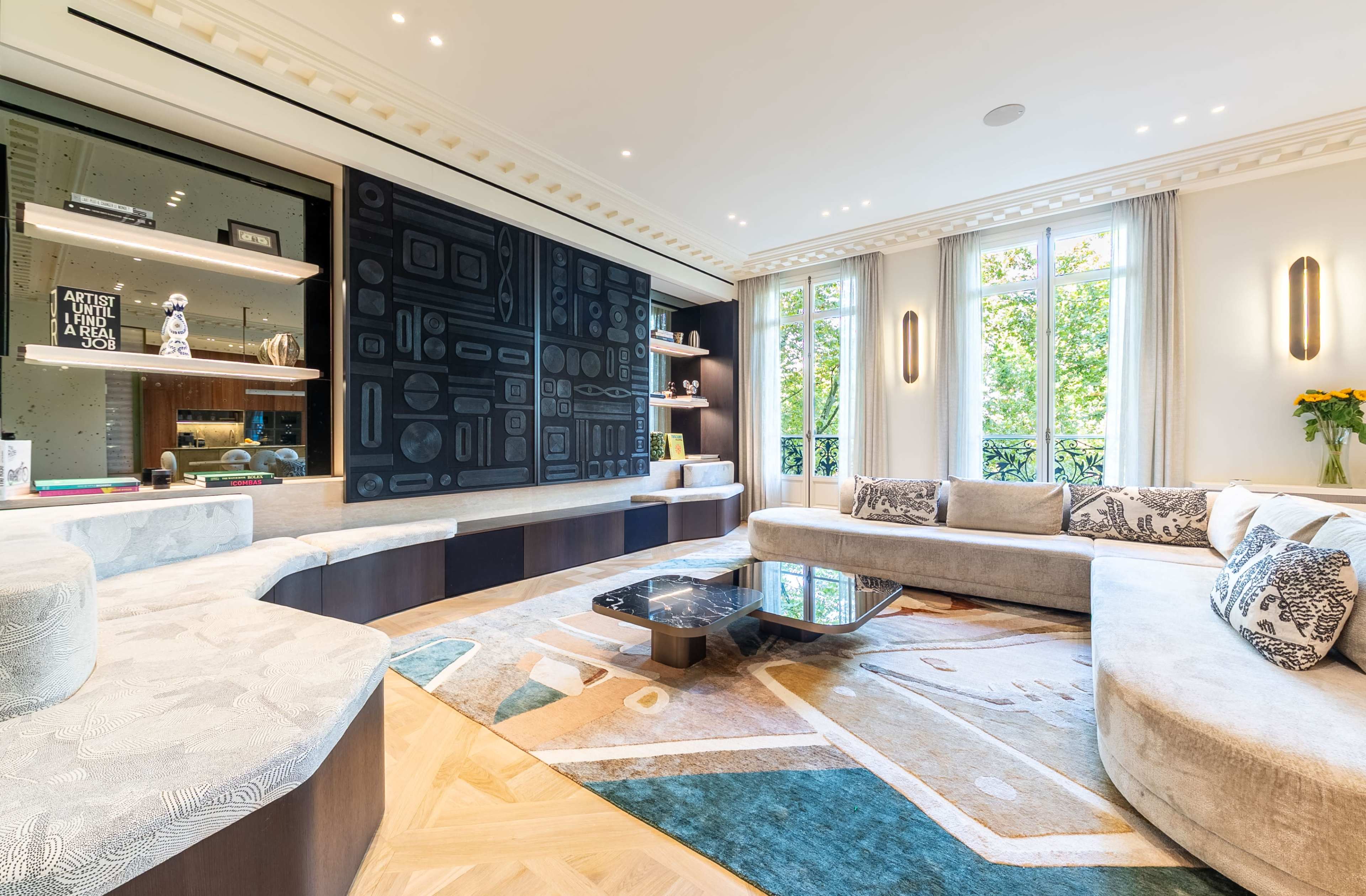 The image shows a modern living room featuring a curved sectional sofa, a coffee table, decorative artwork, and large windows with a view of greenery outside.