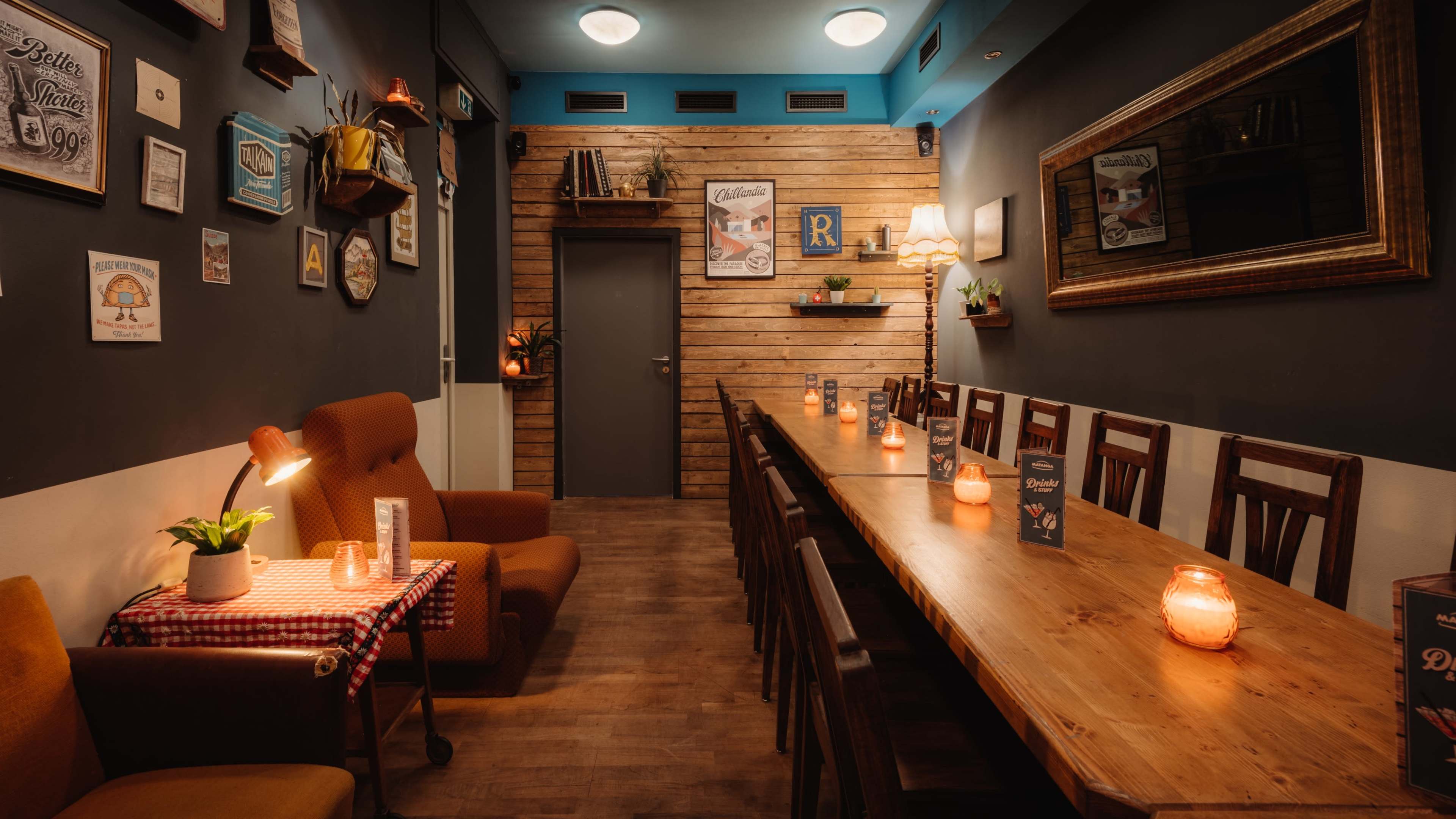 A cozy restaurant interior featuring wooden walls, a long dining table with candles, and a mix of seating arrangements including a sofa and chairs.