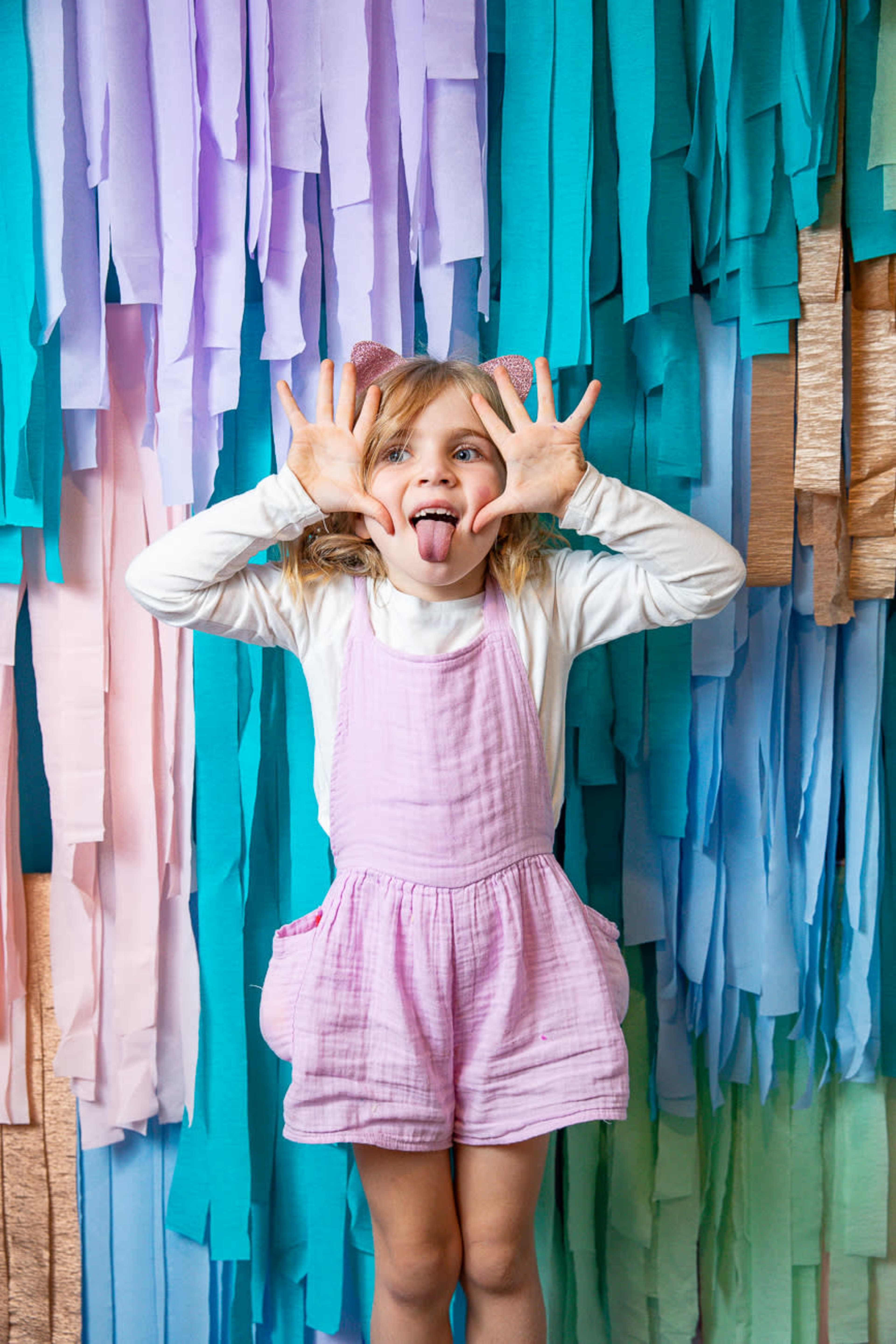 A girl with pigtails and a pink outfit playfully sticks out her tongue while making hand gestures in front of a colorful backdrop of fabric strips.