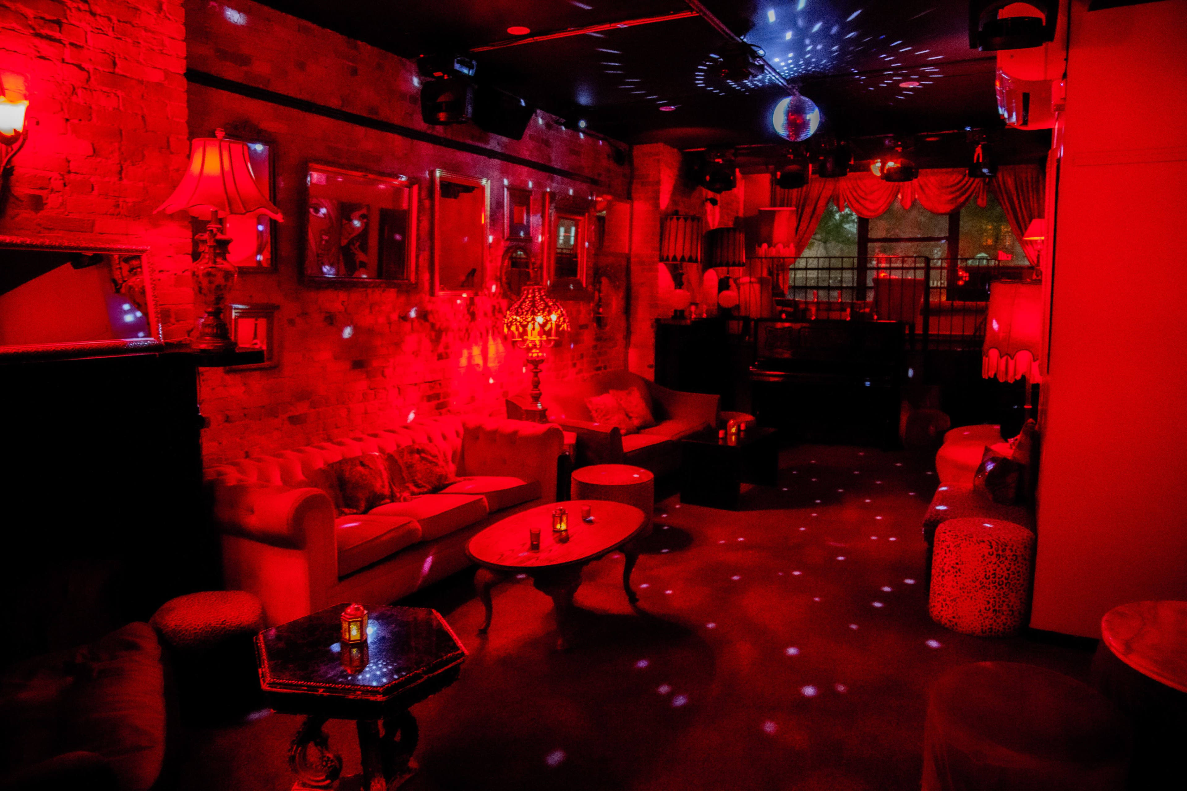 The image shows an illuminated lounge filled with red lighting, featuring a vintage sofa, tables, and decor, along with a disco ball reflecting light across the room.