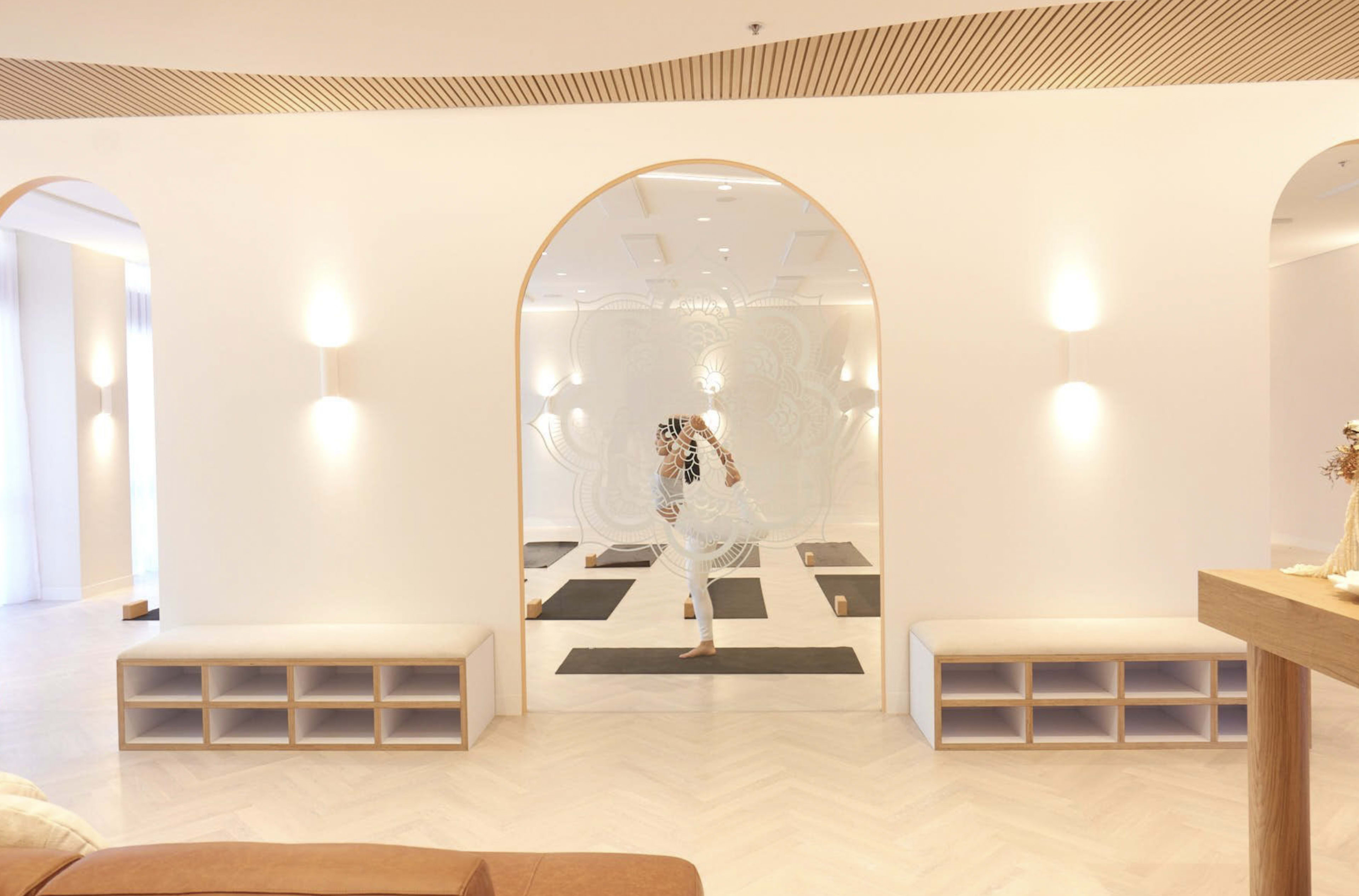 A person is practicing yoga in a minimalist studio with wooden benches and mirrored walls.