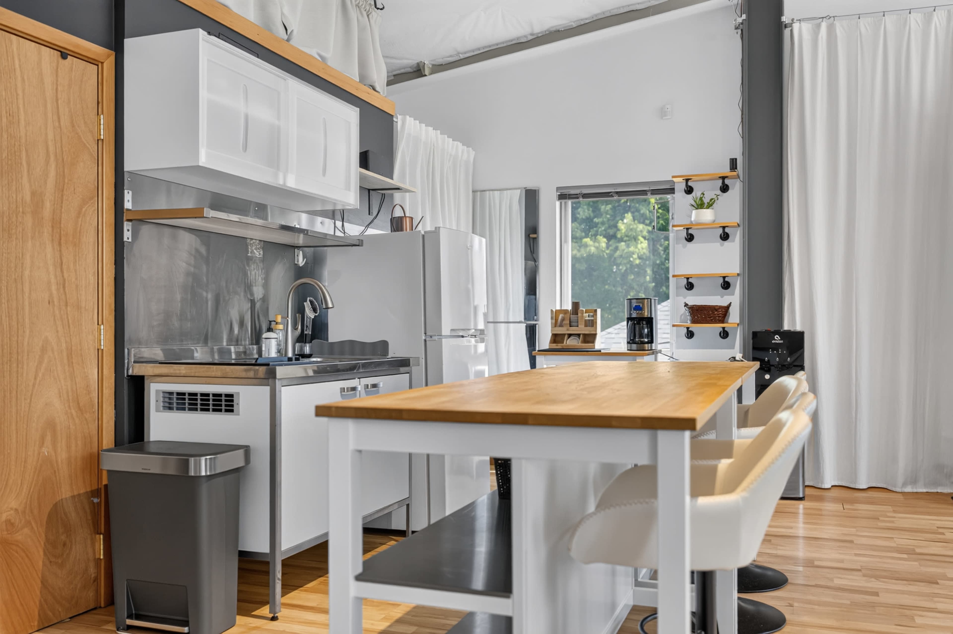 The image shows a modern kitchen area with a wooden table, white cabinetry, stainless steel appliances, and large windows draped with white curtains.