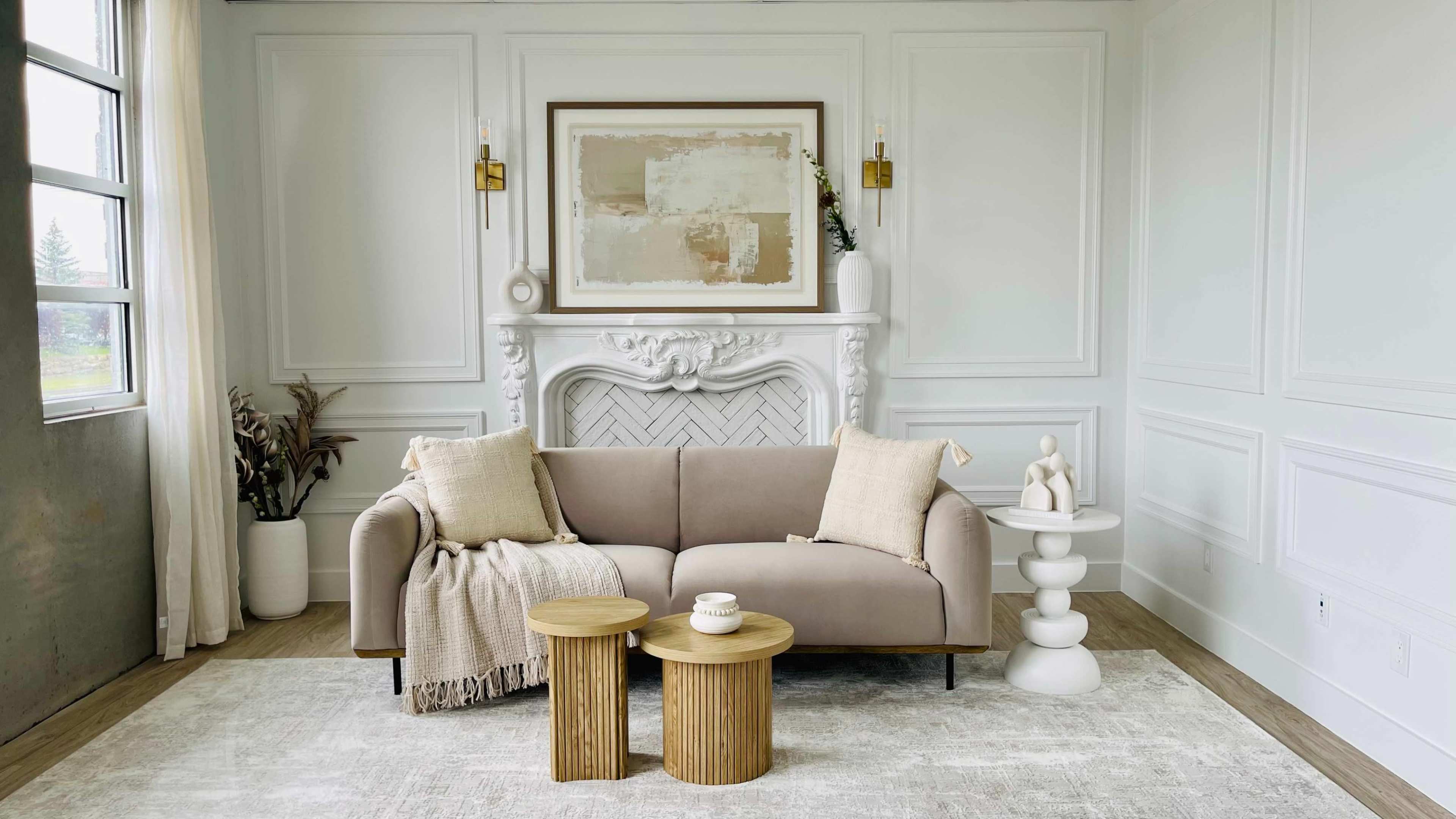The image depicts a modern living room featuring a beige sofa, two round wooden tables, and a decorative fireplace with wall art above it, all set against a light-colored wall.