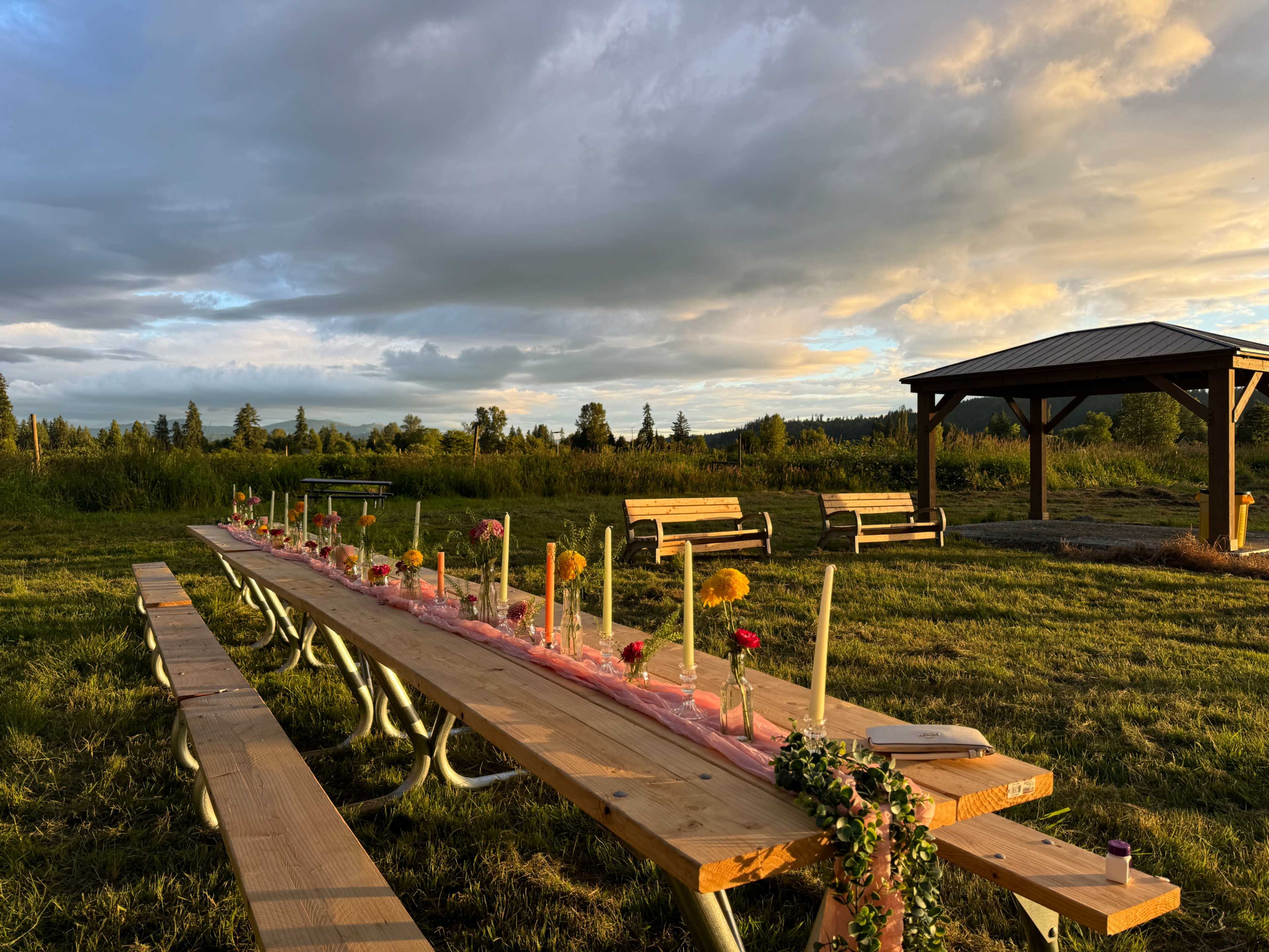 A long wooden table is set with flowers and candles under a cloudy sky in a grassy area beside a shelter and benches.