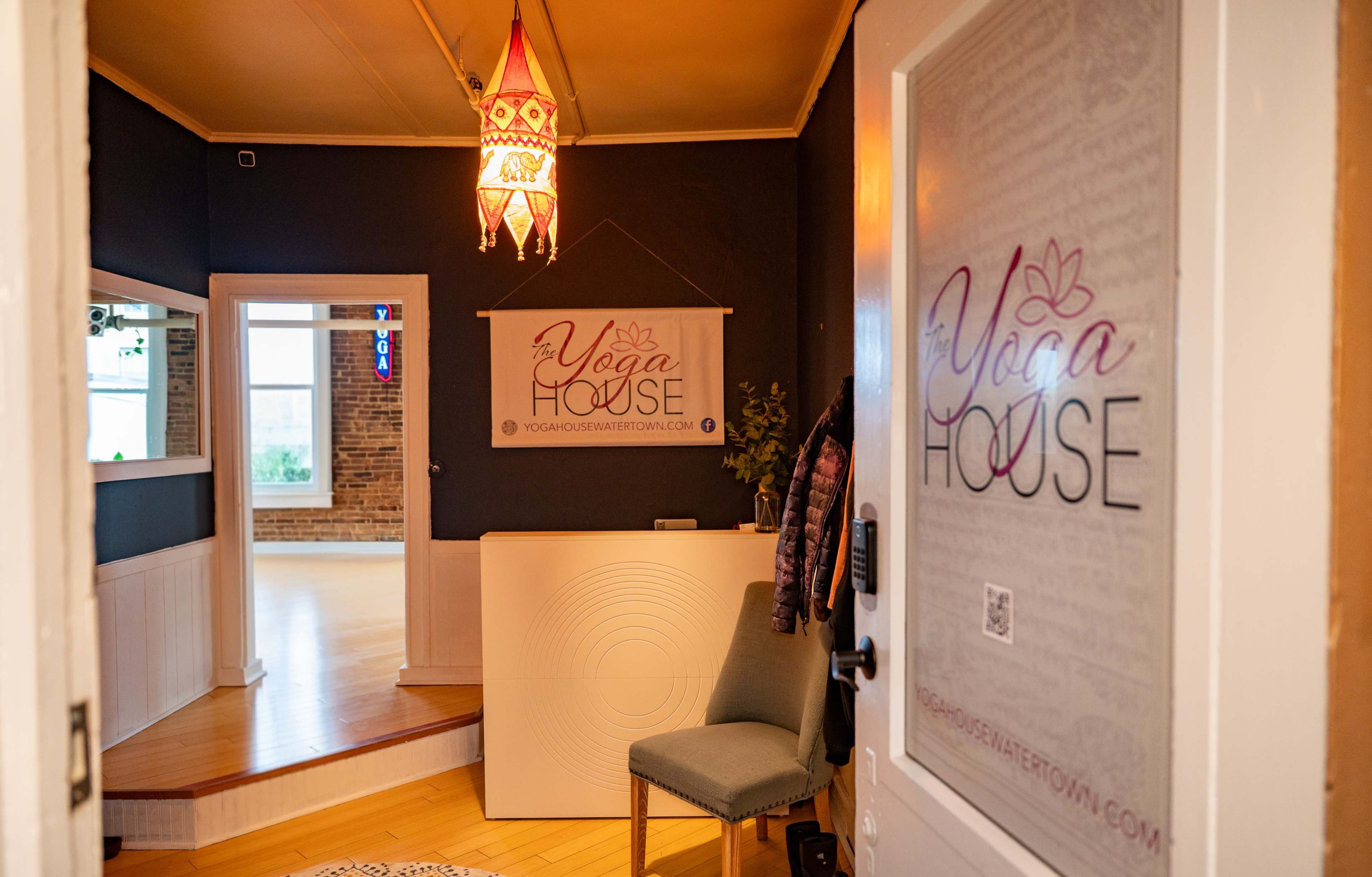 The image shows the entrance of a yoga studio with a welcoming sign and a chair, featuring wooden flooring and a light fixture above.