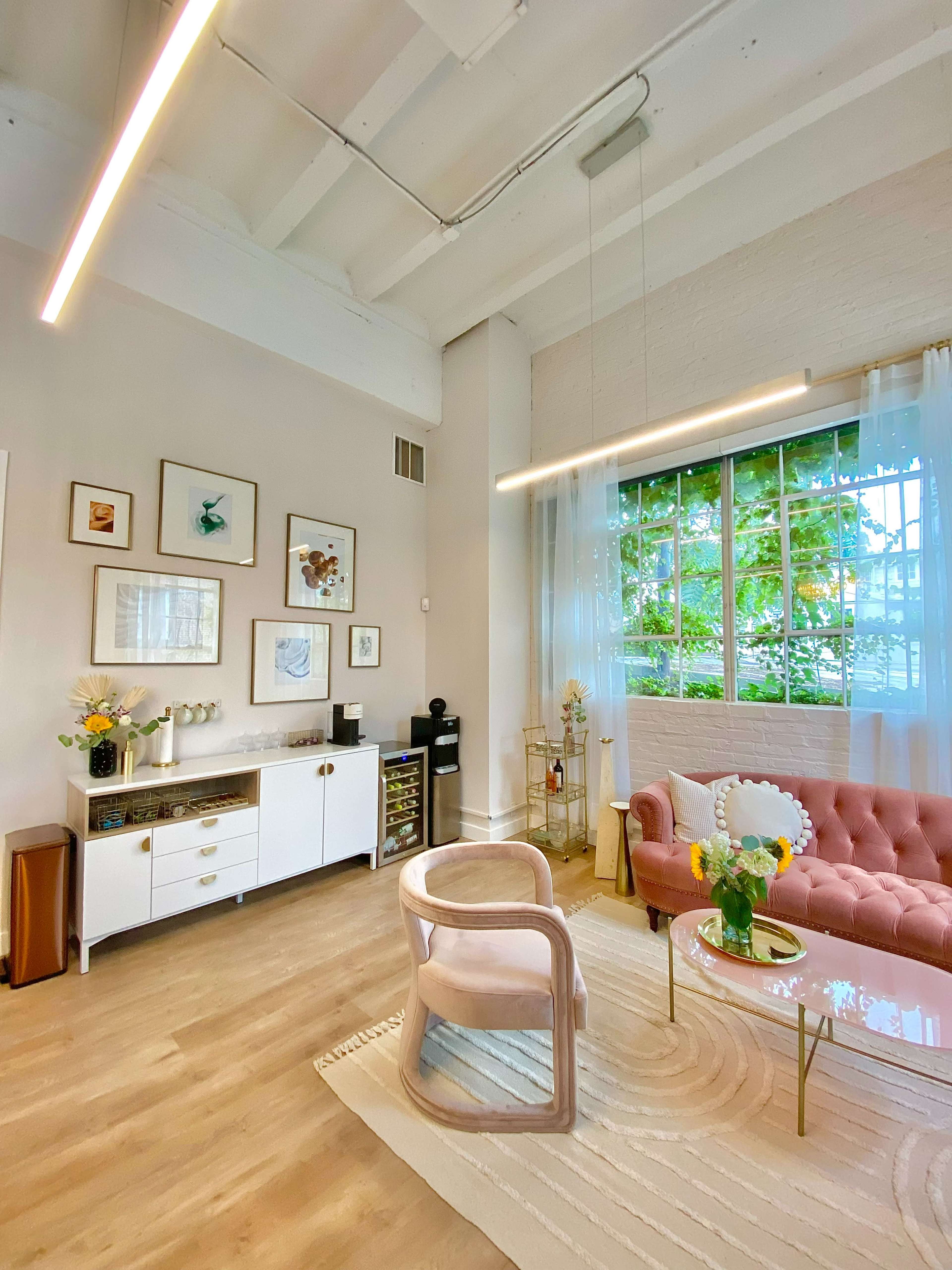 The image shows a bright interior featuring a pink tufted sofa, a glass coffee table with flowers, and a window with sheer curtains allowing natural light to filter in.