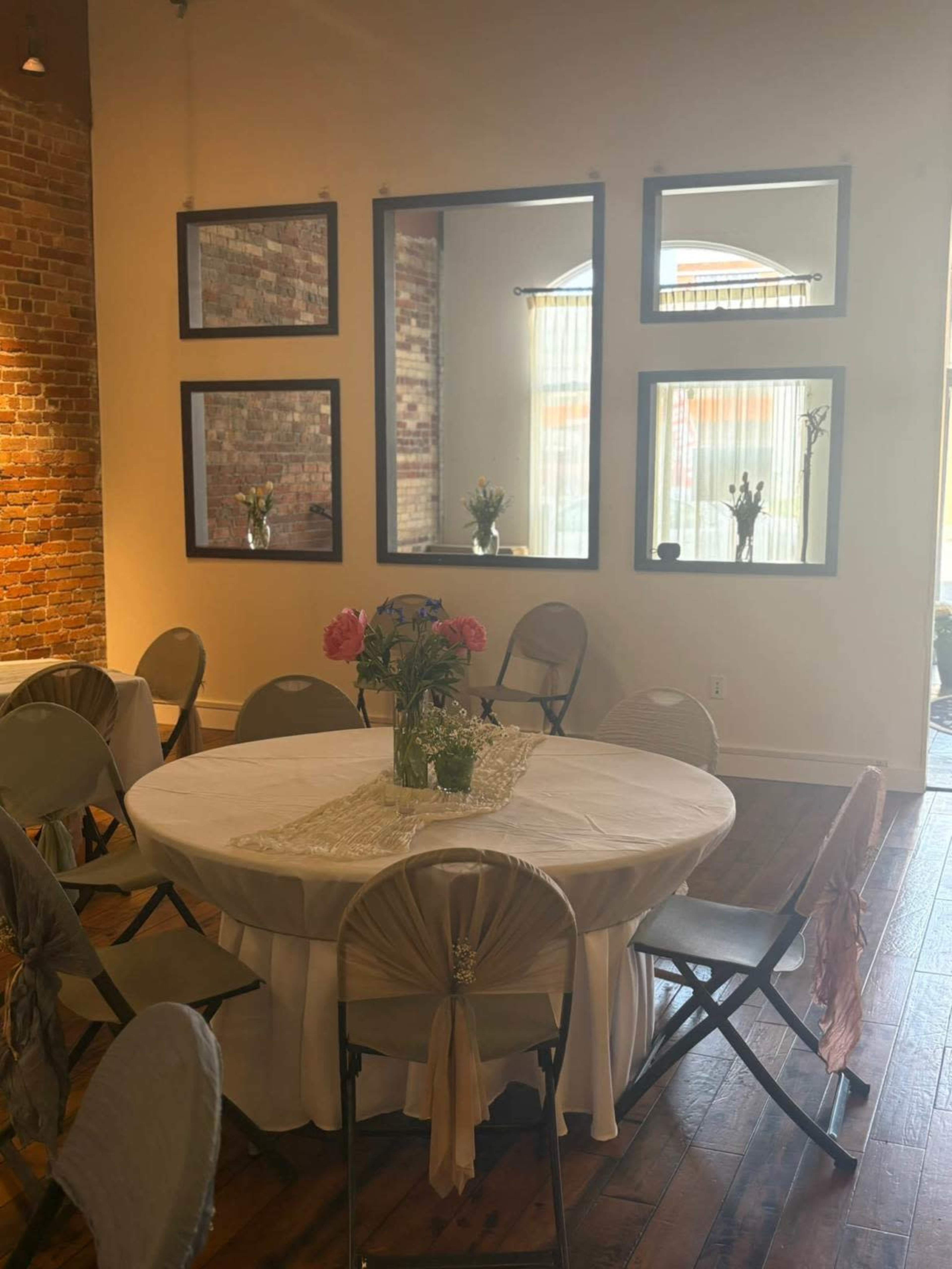 The image shows a decorated dining area with a round table set for an event, surrounded by folding chairs and featuring brick walls and framed mirrors.