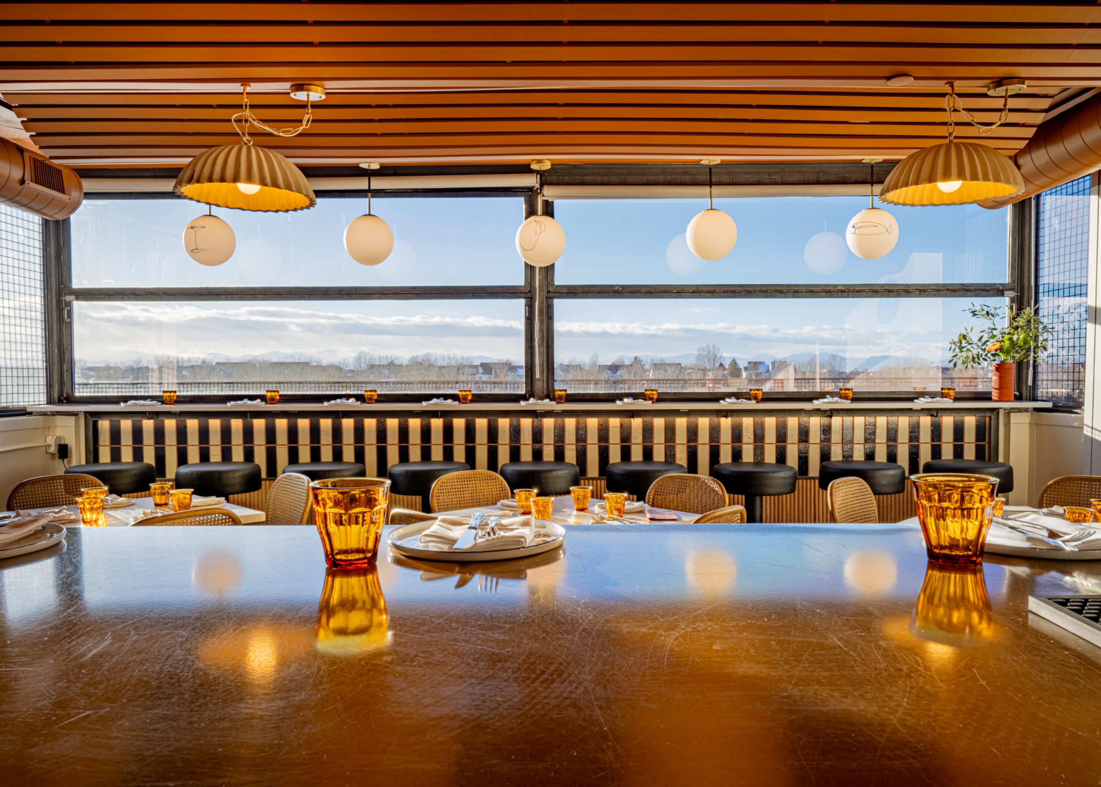 The image shows a modern restaurant interior with a long wooden table set for dining, surrounded by a row of chairs, and large windows offering a view of the outside.