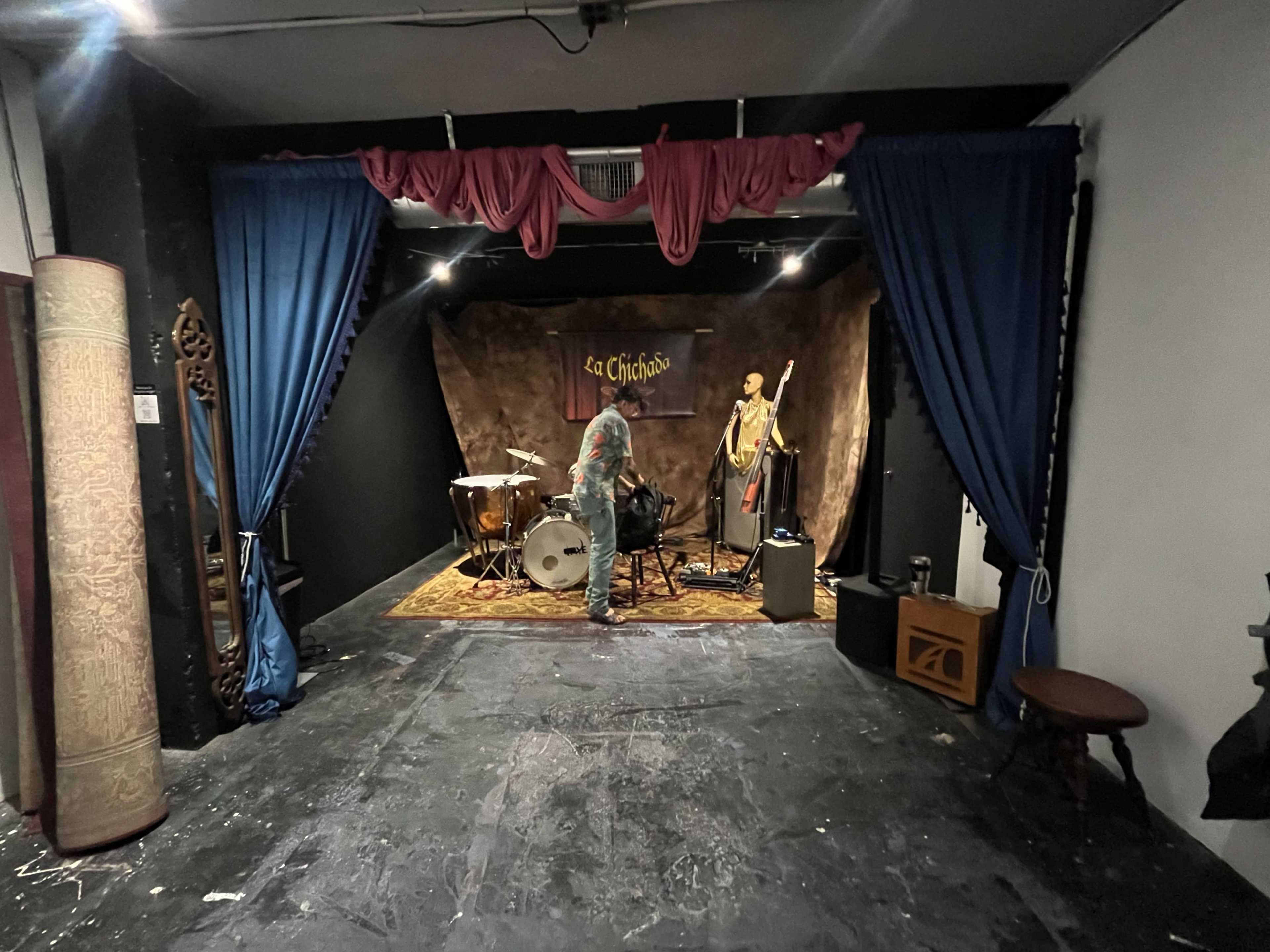 The image shows a small stage set up for a performance, complete with a drum set, microphone, and backdrops adorned with curtains and a banner that reads "La Chichón."