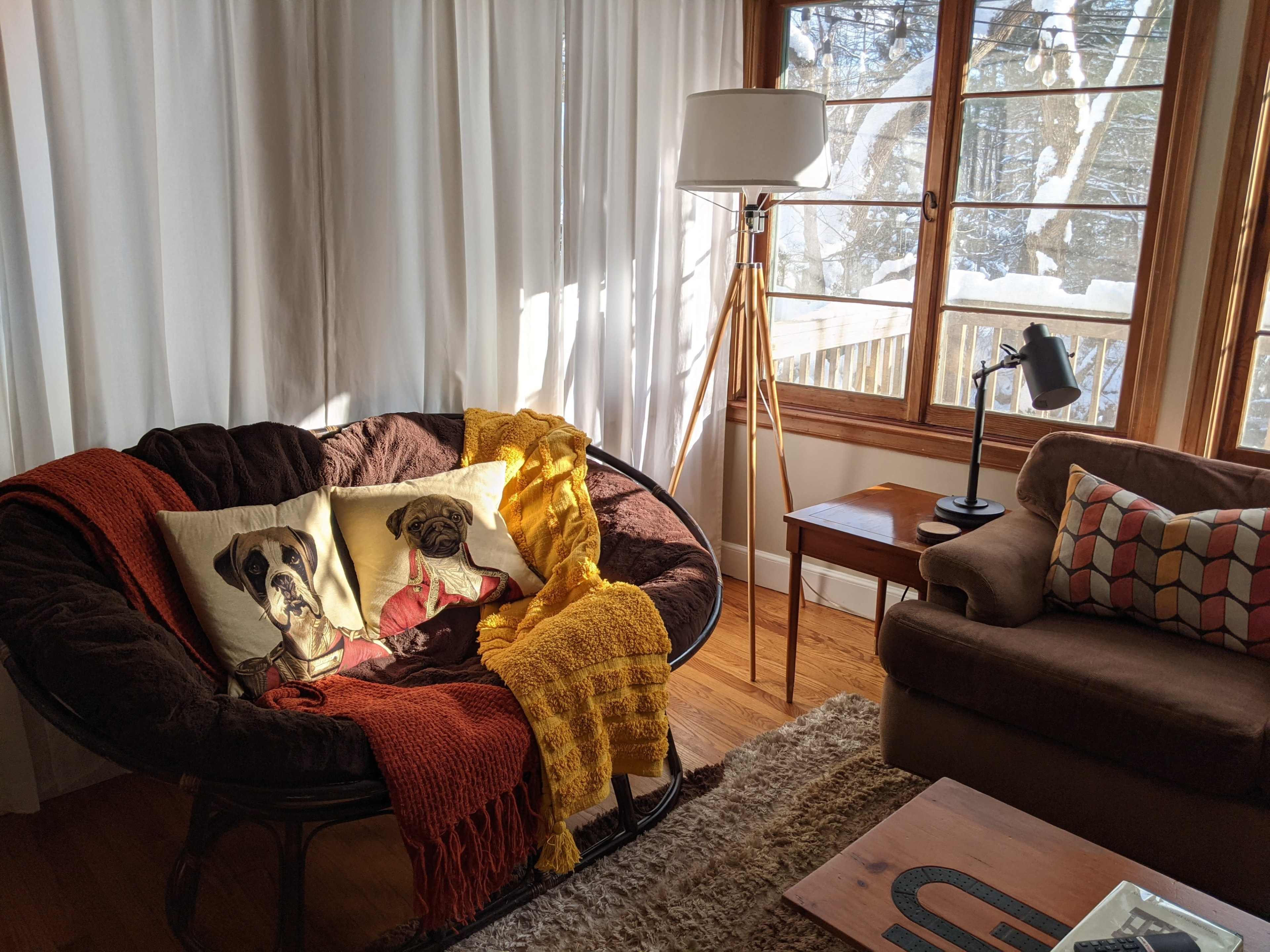 A cozy living room features a circular chair adorned with dog-themed pillows, a yellow throw, and a wooden lamp beside a window with sheer curtains.