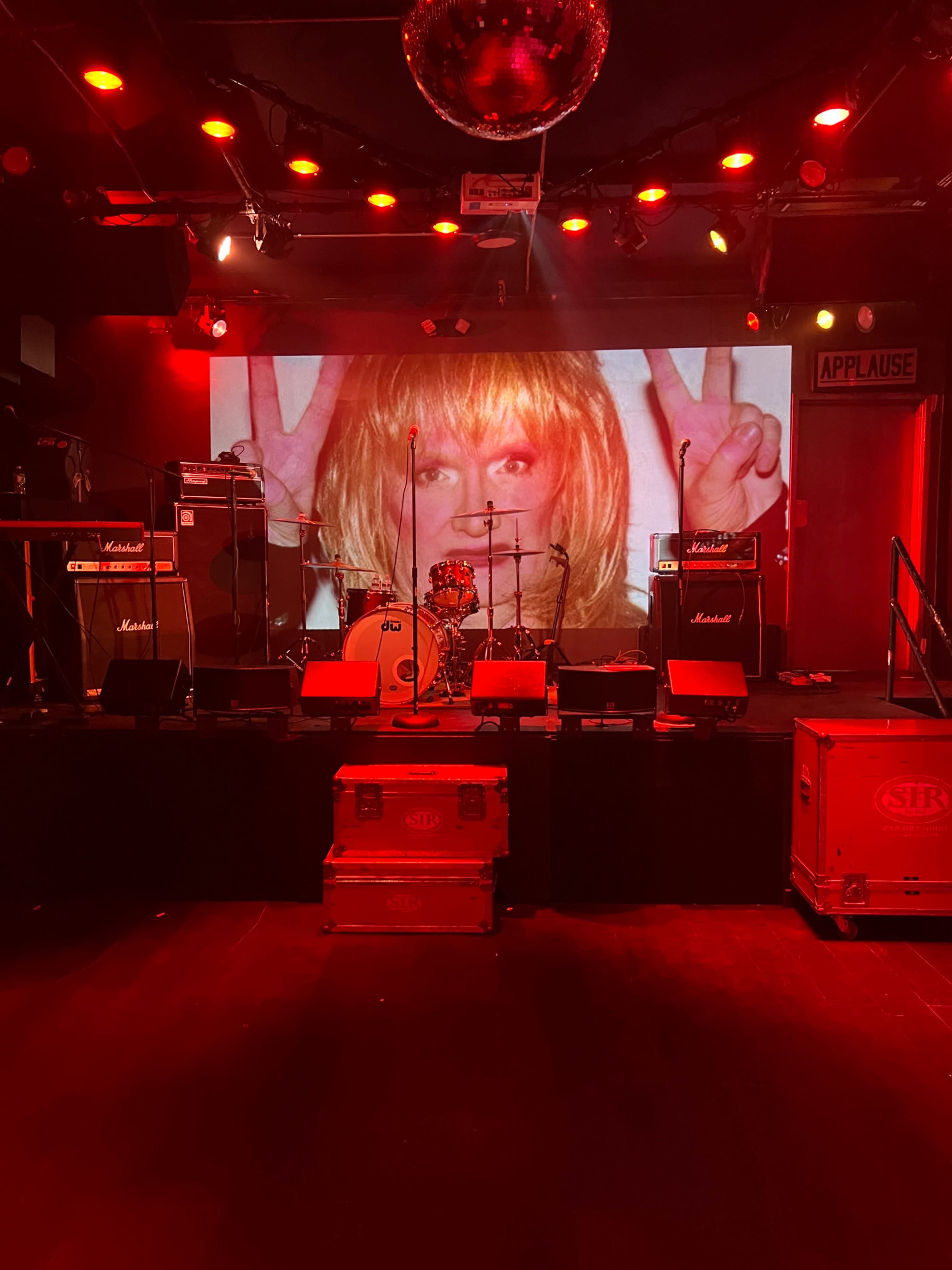 A stage is set with amplifiers and a drum set in front of a large backdrop displaying an image of a woman with blonde hair making a peace sign.