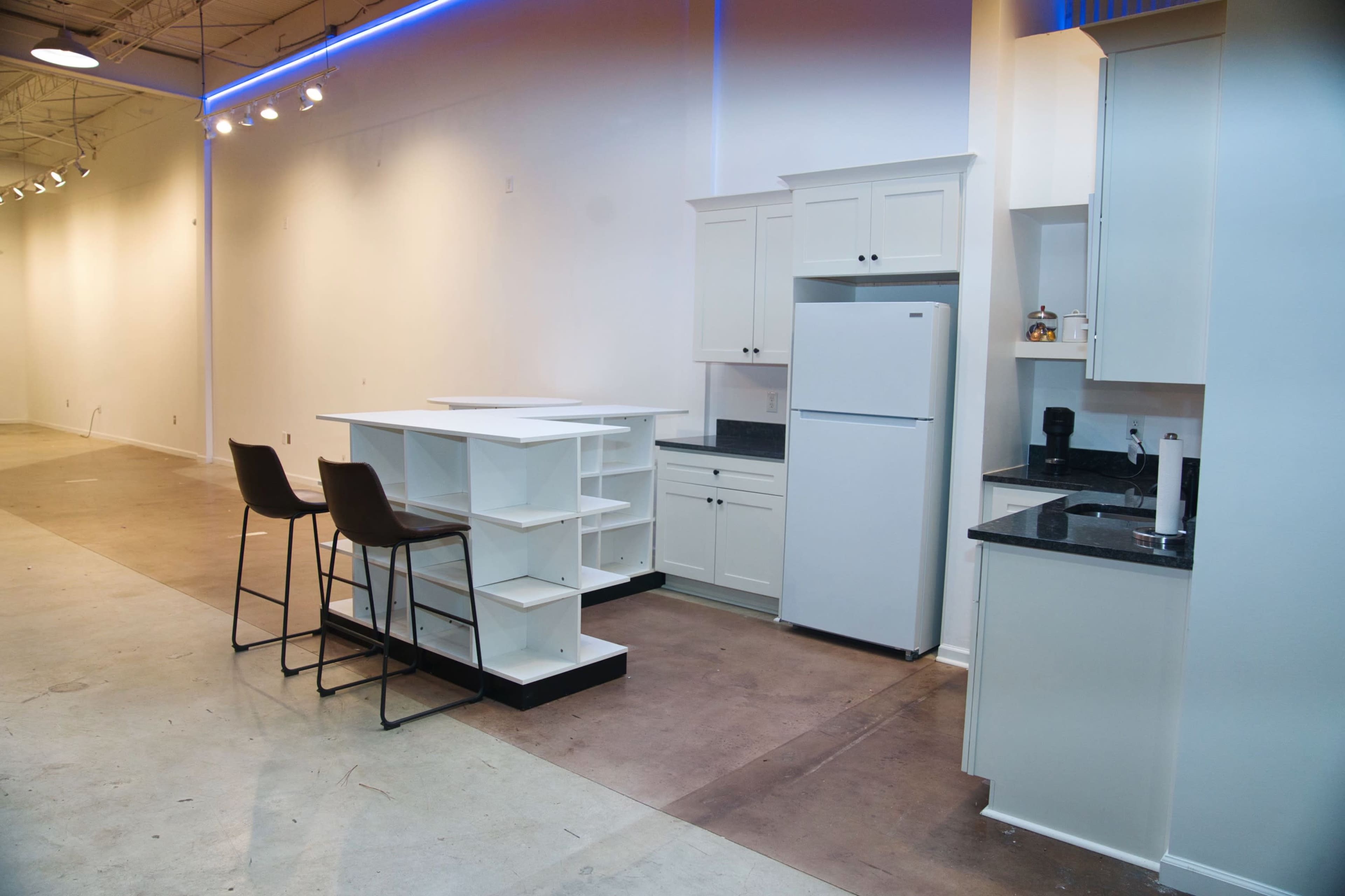 A modern kitchen setup featuring a white refrigerator, cabinets, and an island with two bar stools in an empty, well-lit space.