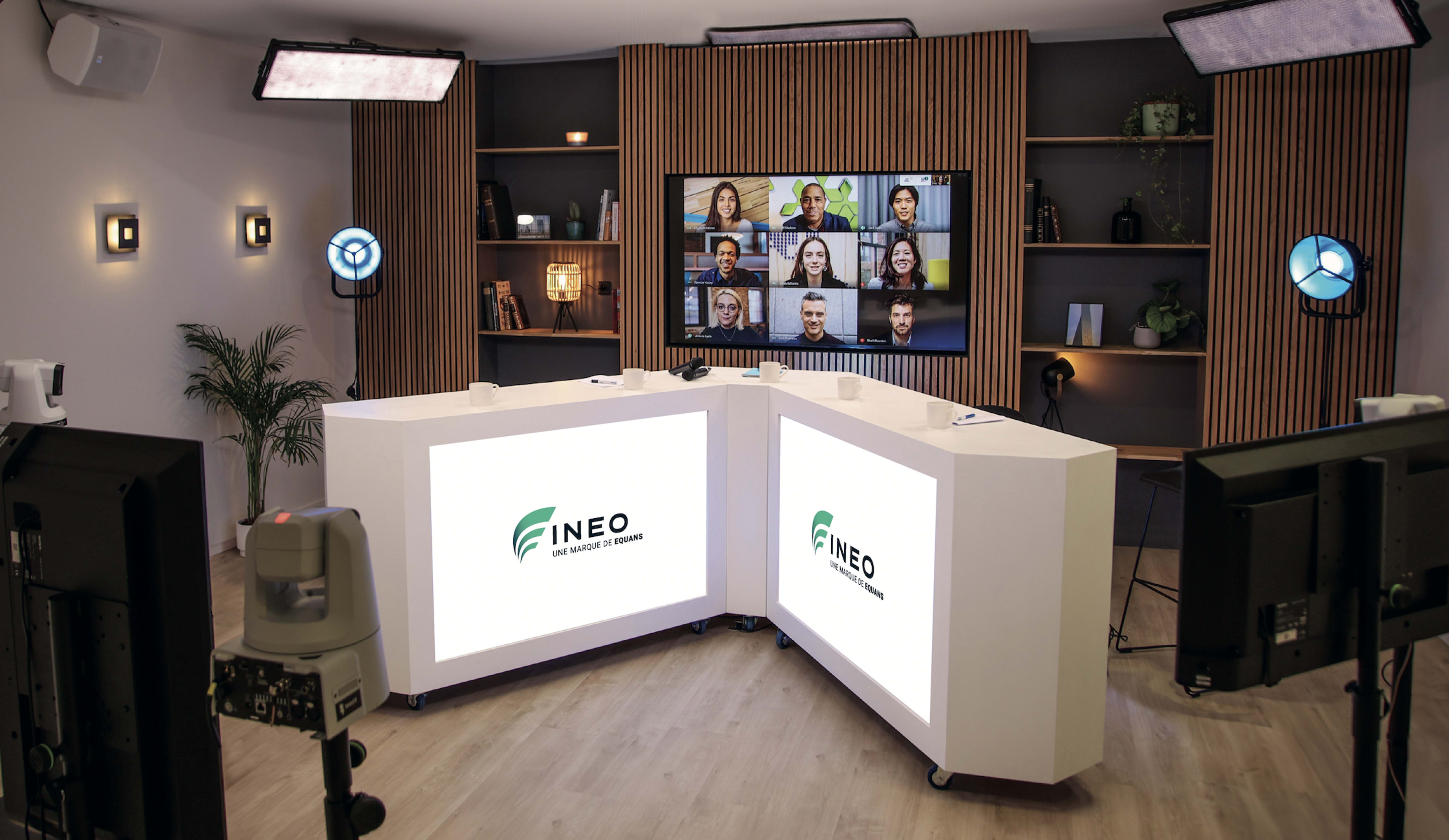 The image shows a modern studio setup with two large white curved tables displaying the logo "FINEO," surrounded by cameras and lights, and a screen featuring a grid of video call participants.