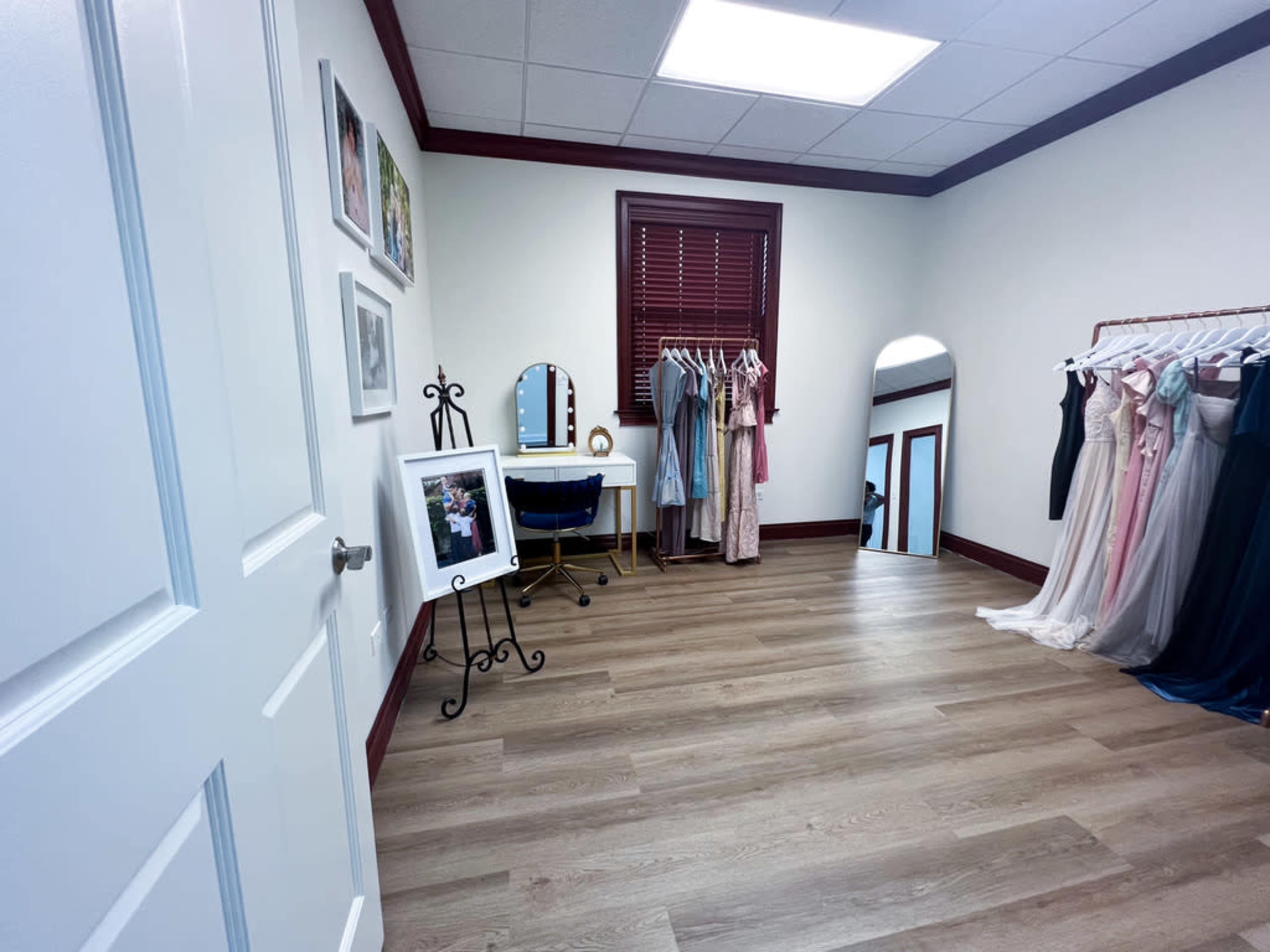 A room with wooden flooring, a mirror, a small table, and several dresses displayed on racks, along with framed photos on the walls.