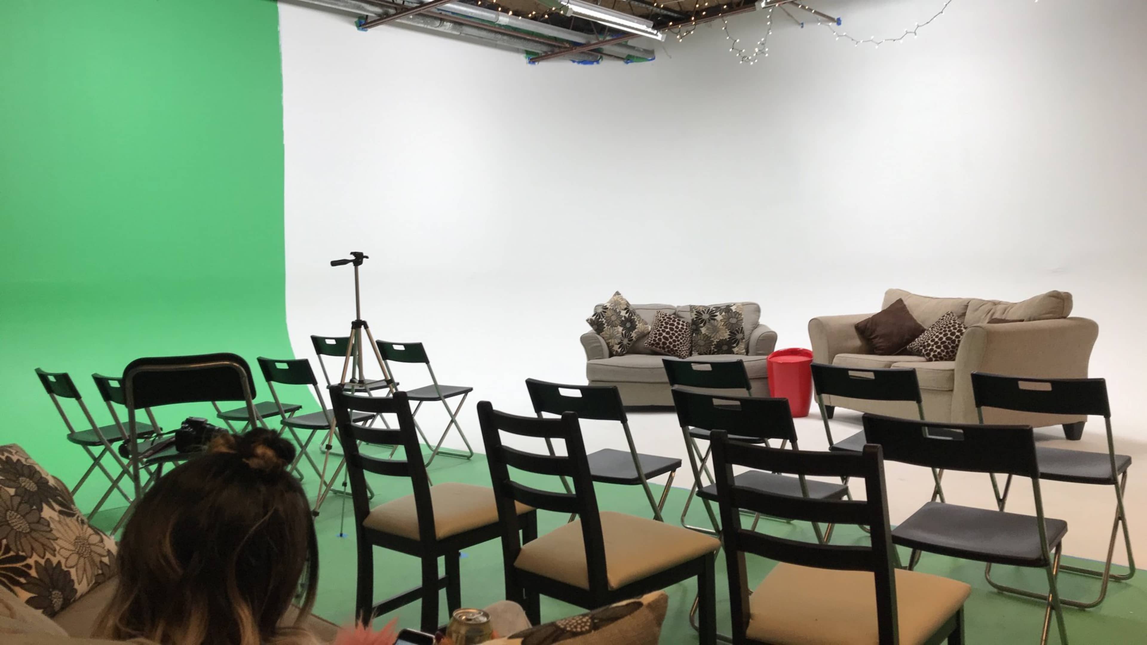 A studio setup with two sofas, several black chairs arranged in rows, a microphone stand, and a green-screen wall on one side.