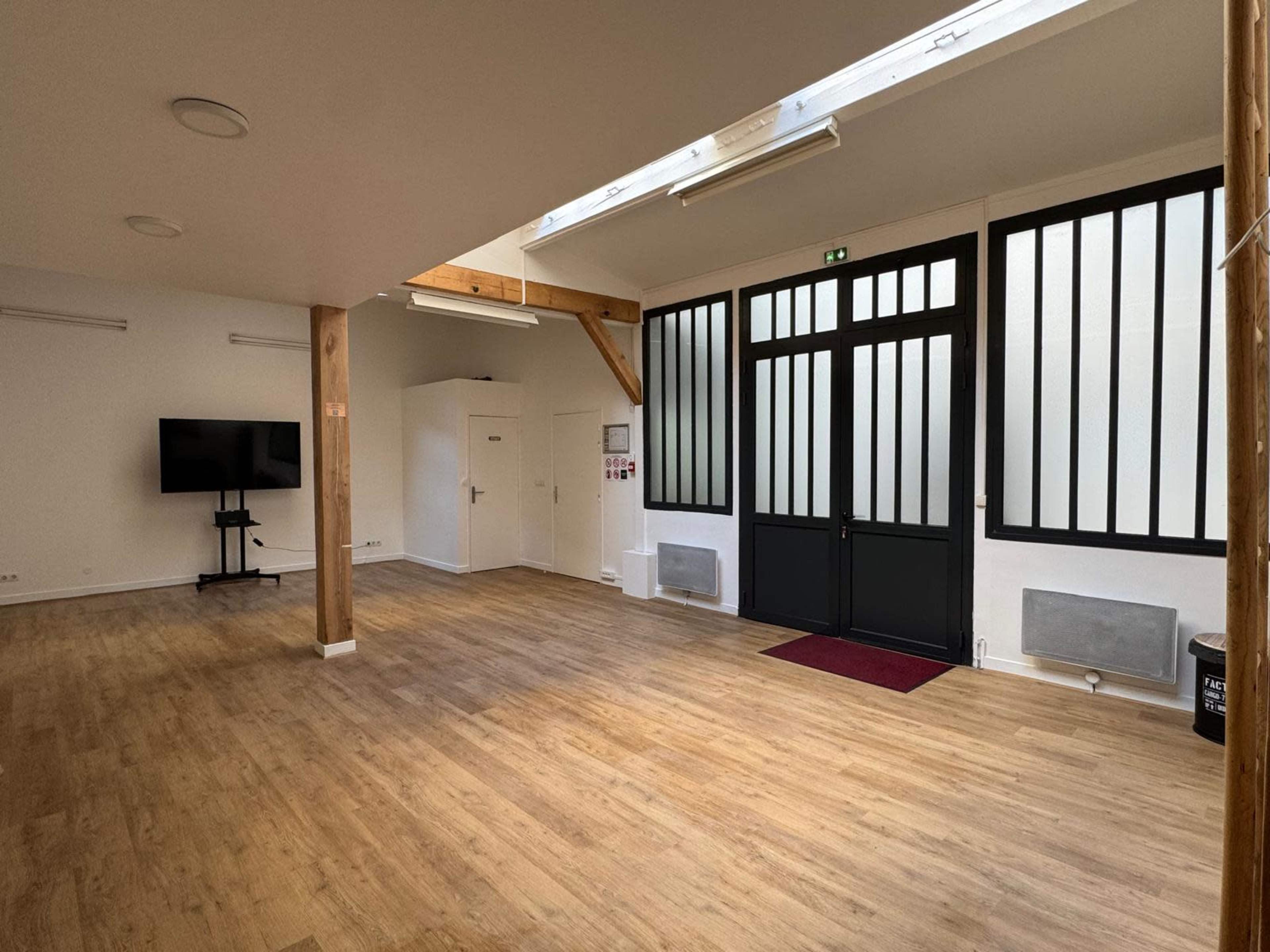 The image shows a spacious, well-lit interior room with wooden floors, large windows, and a television mounted on the wall.