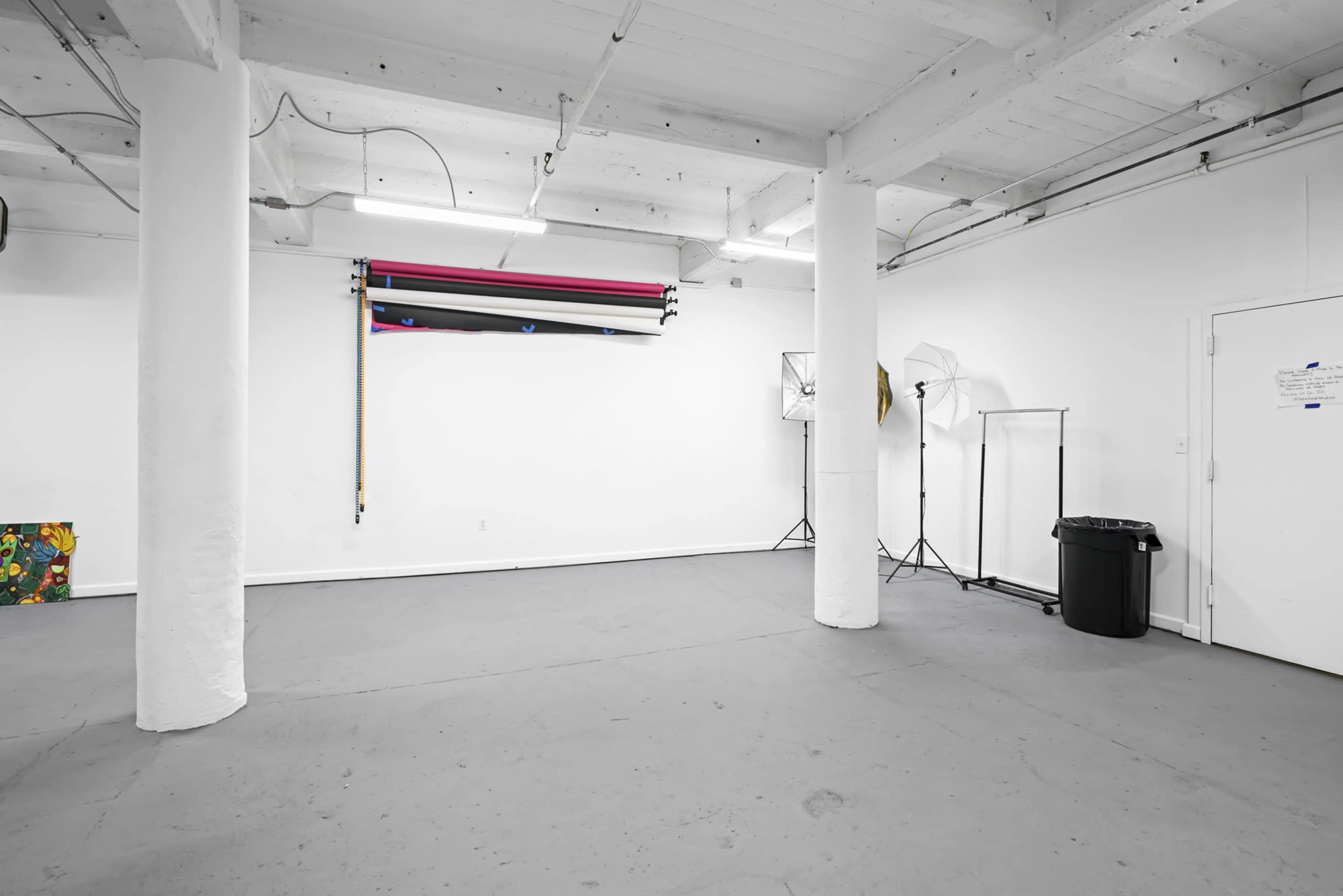 A spacious, empty studio with white walls, a gray floor, and various photography equipment, including backdrop rolls and lighting stands.
