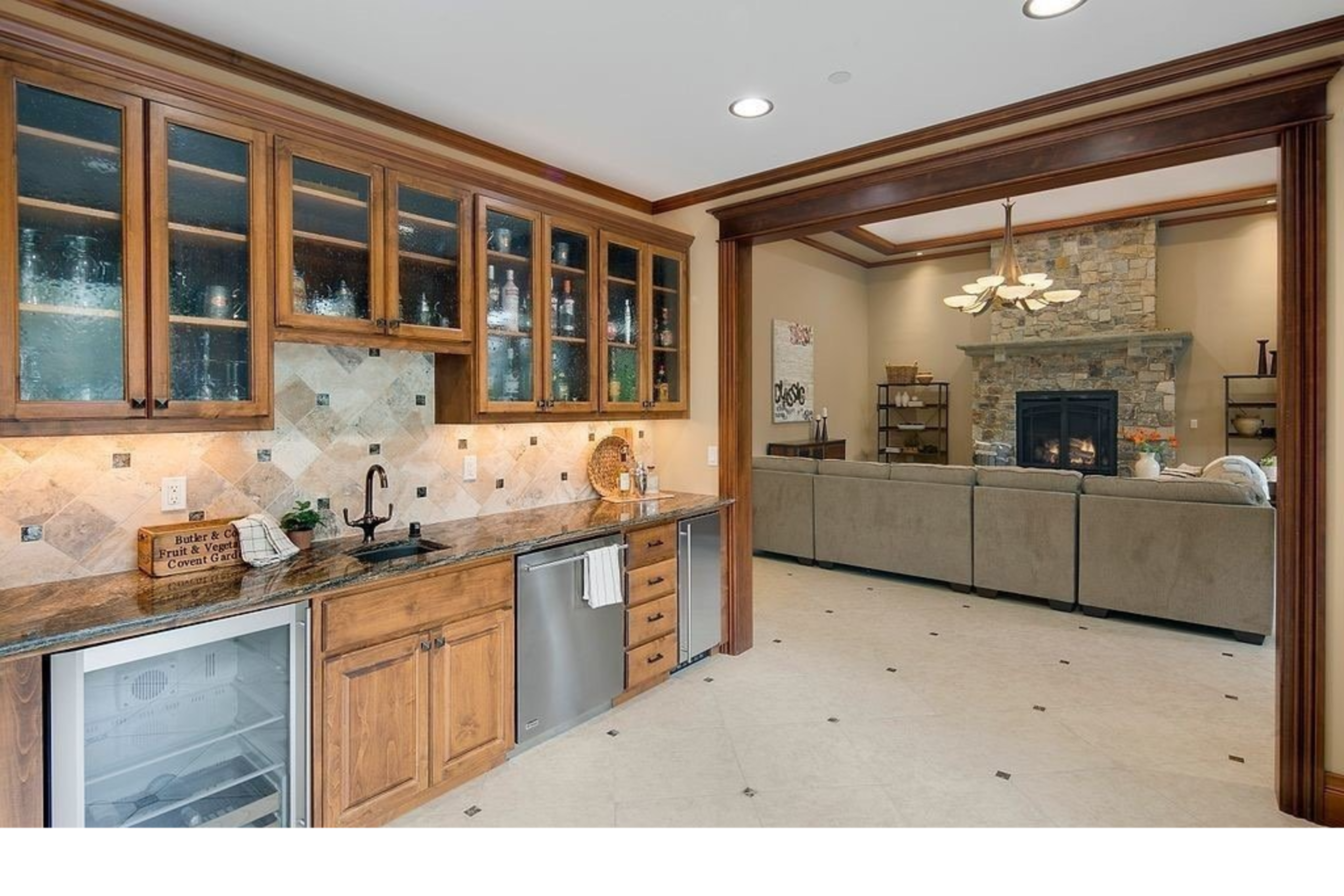 The image shows a kitchen with wooden cabinetry, a stone backsplash, and a view of a living area featuring a beige sofa and a stone fireplace.