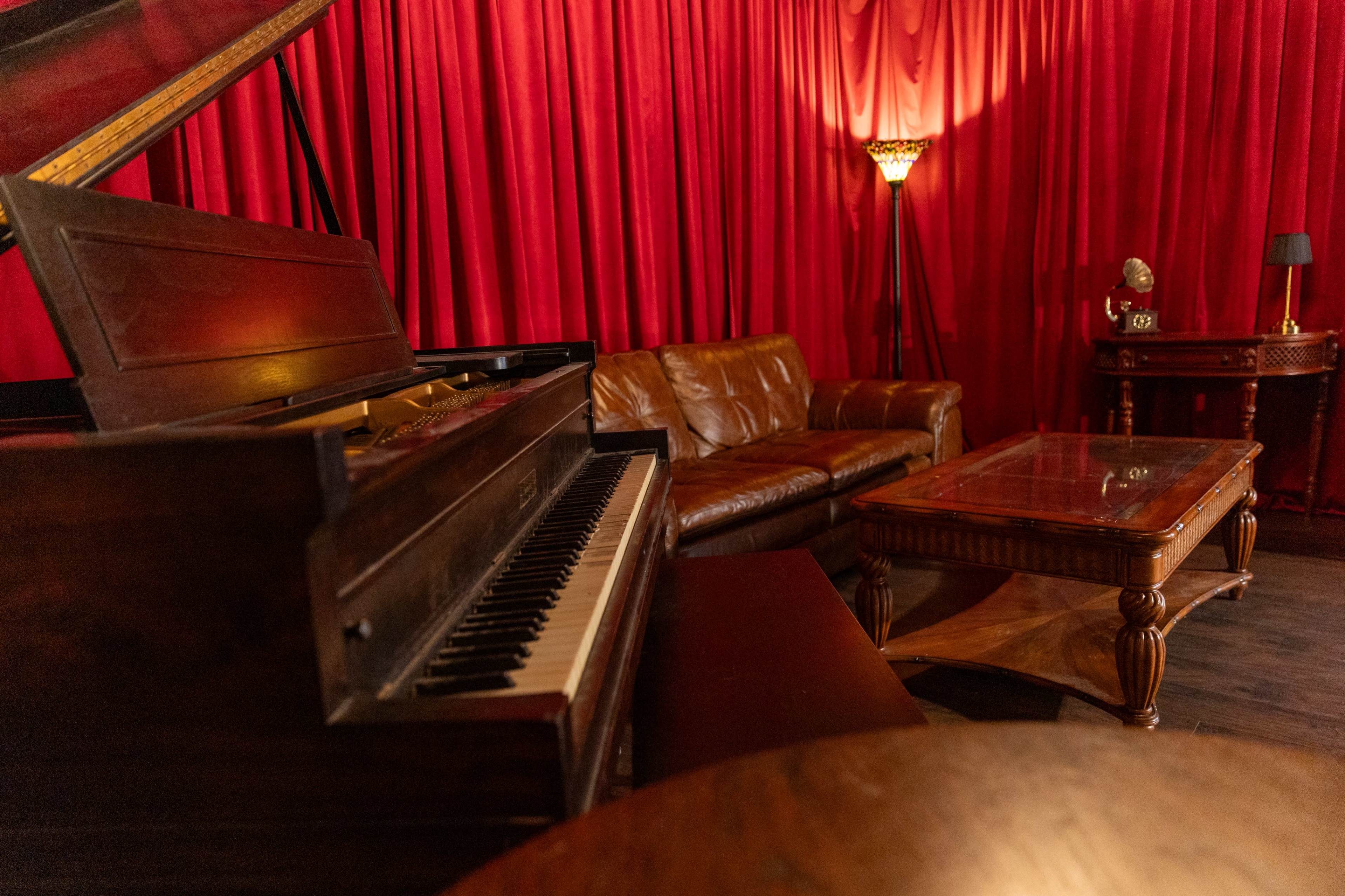A vintage piano is positioned next to a brown leather couch in a room with red curtains and warm lighting.