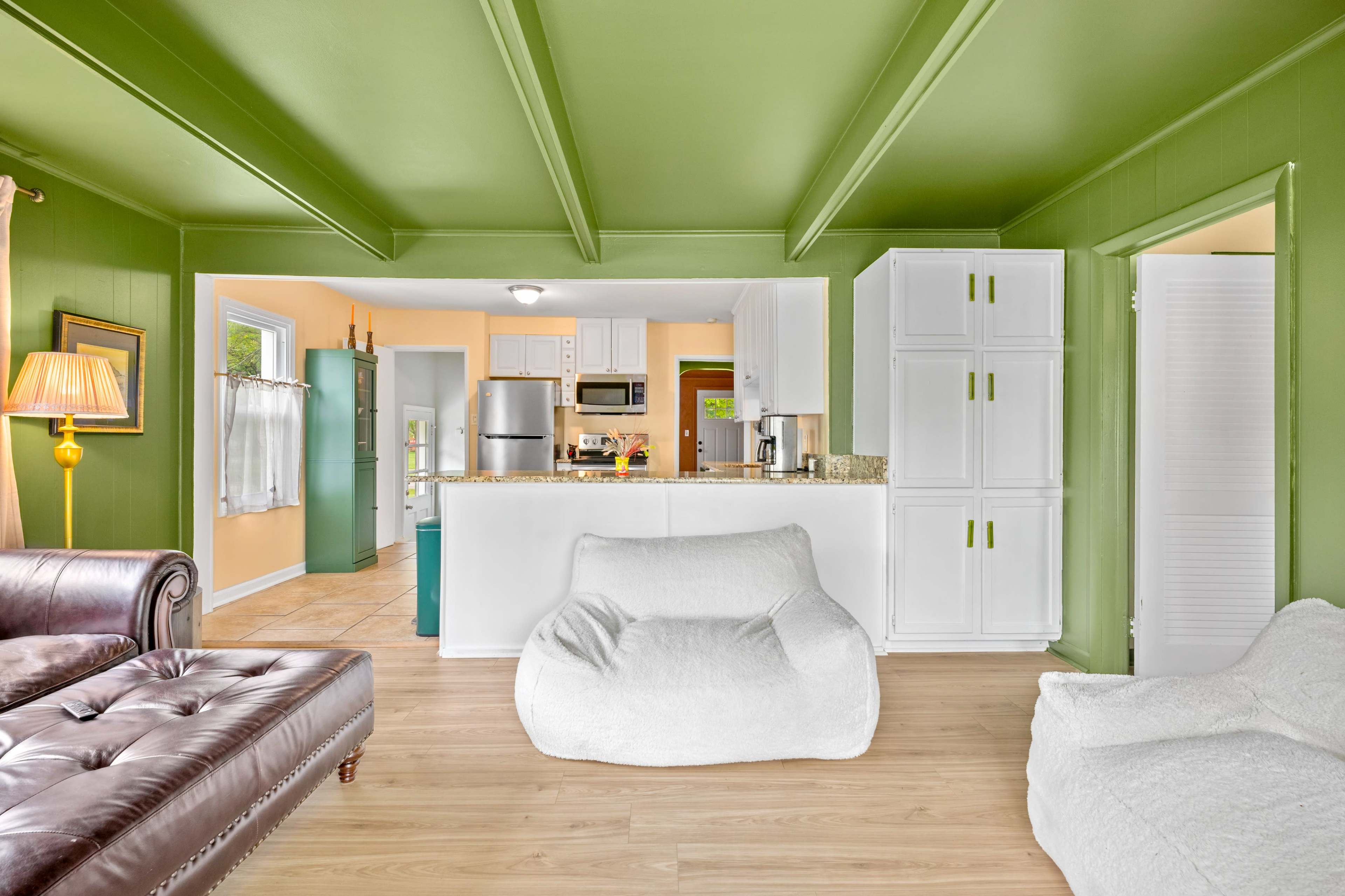 The image shows a living room with green walls, a brown leather couch, two white bean bag chairs, and an open kitchen area in the background.