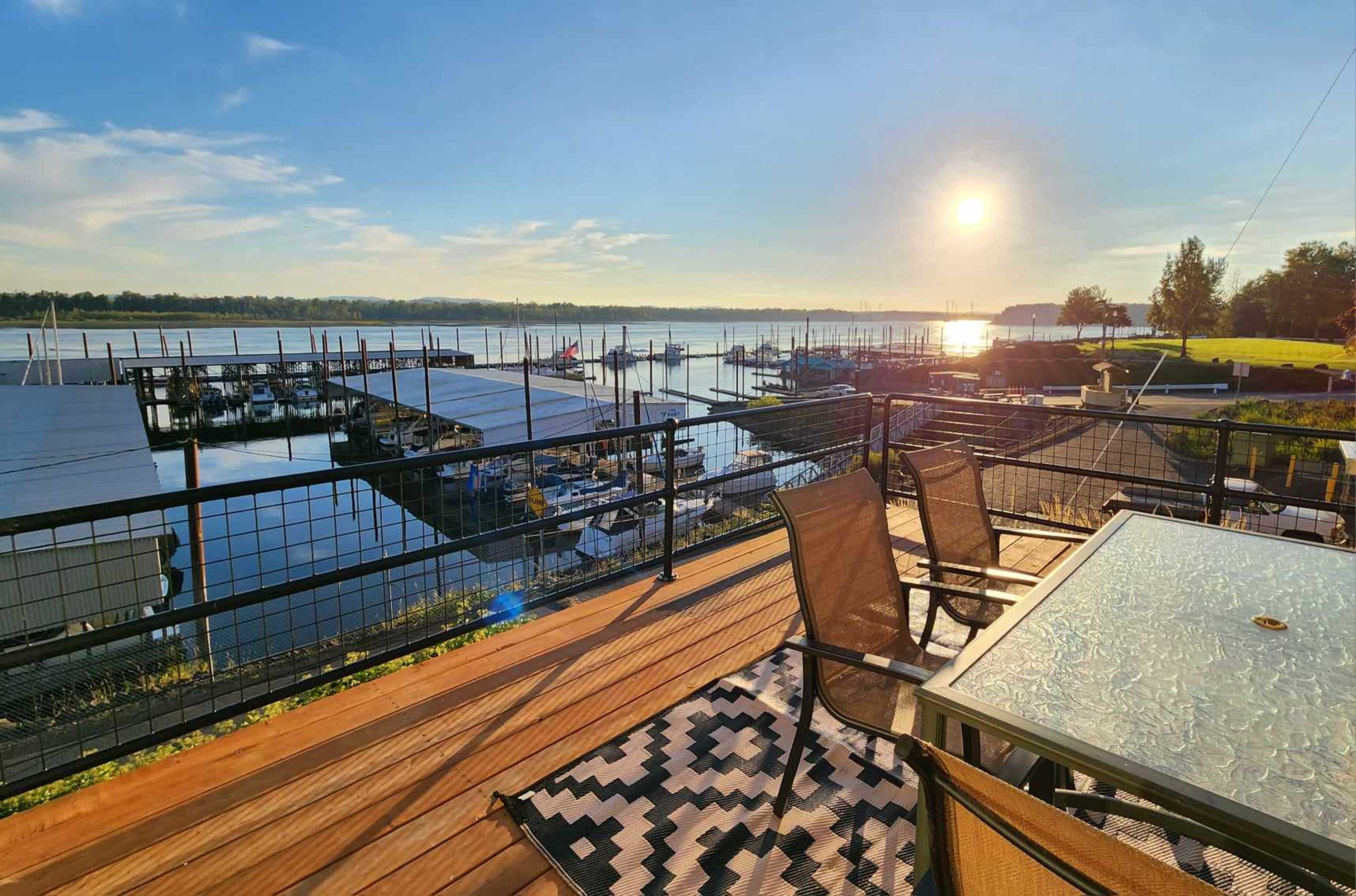 A marina at sunrise with boats docked, a wooden deck with a table and chairs overlooking the water.