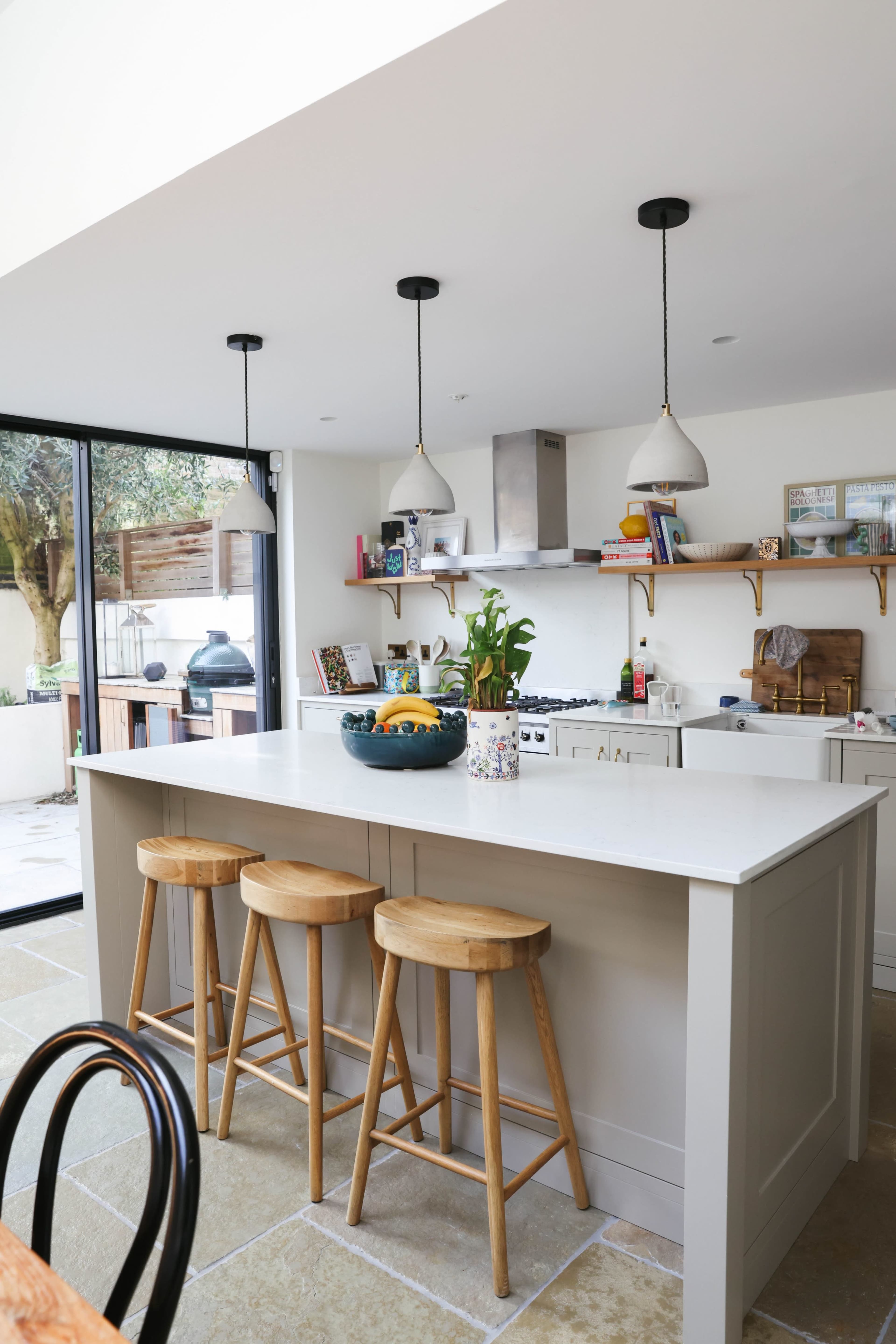 The image shows a modern kitchen featuring a large island with three wooden stools, pendant lights above, and a variety of kitchen appliances and decor along the walls.