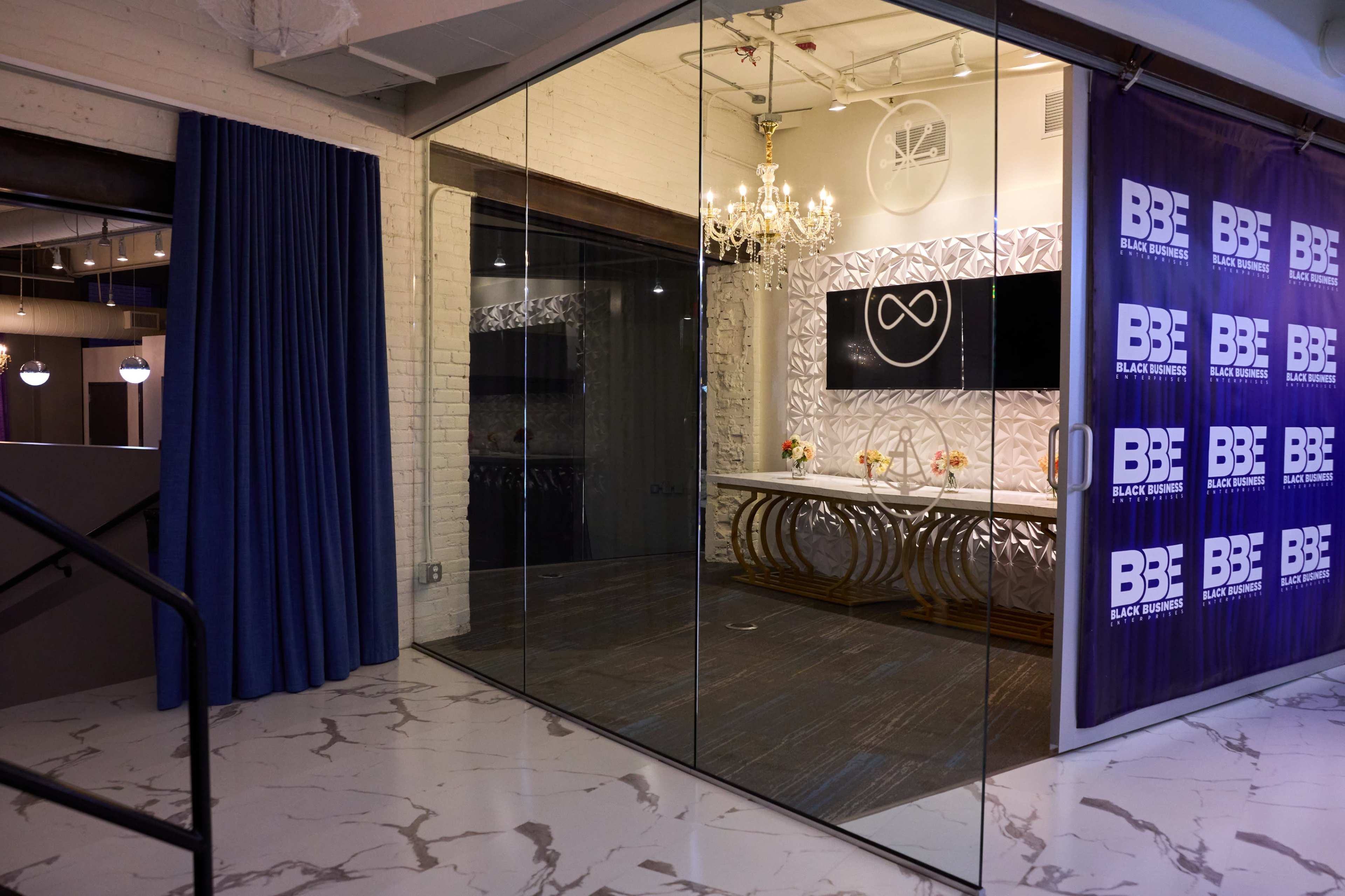 The image shows a modern office space with a glass entrance featuring a decorative wall, a chandelier, and branding elements on a purple banner.