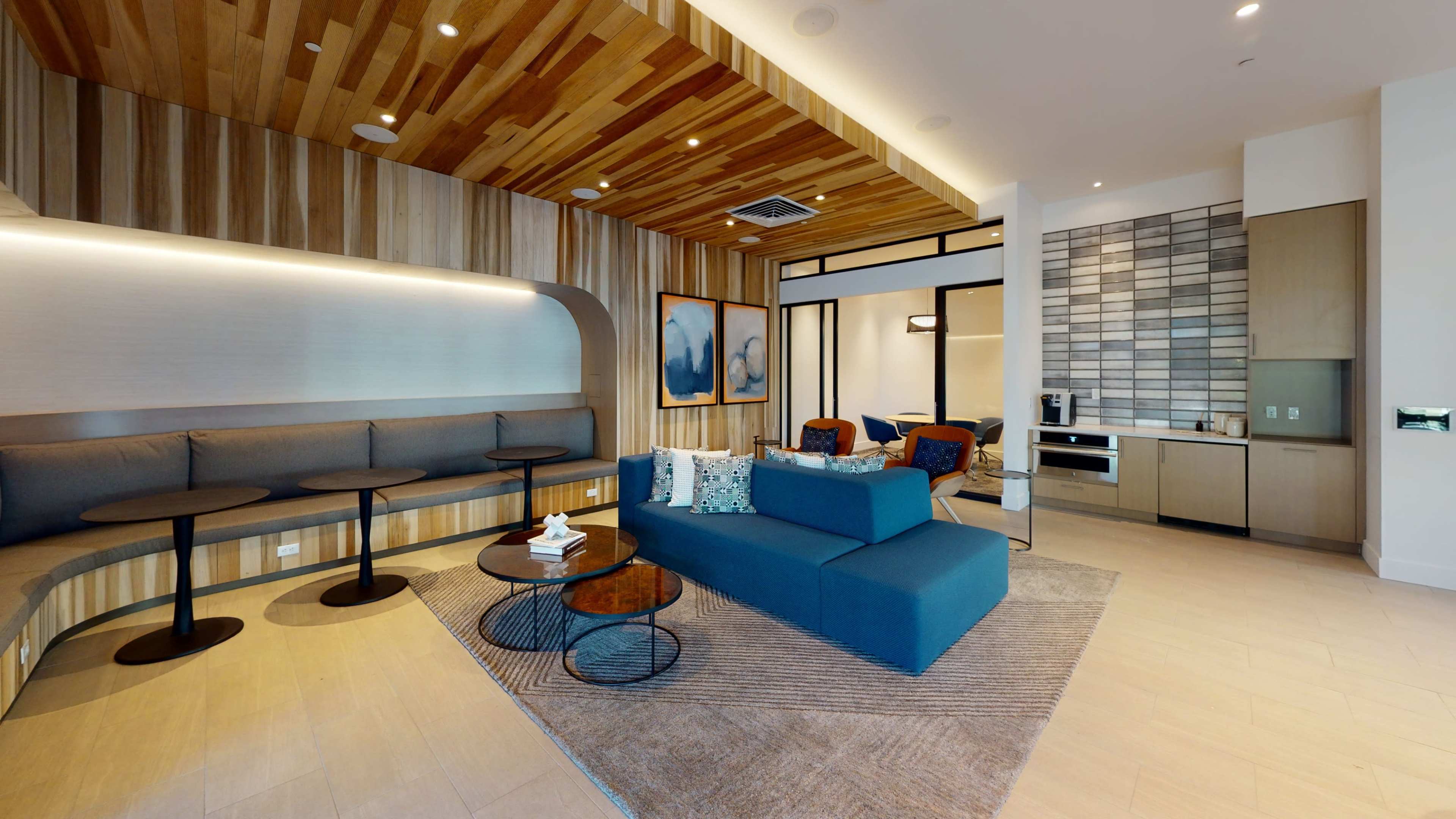The image shows a modern lounge area featuring a blue sectional sofa, round tables, and a kitchenette against a backdrop of wooden paneling and decorative artwork.