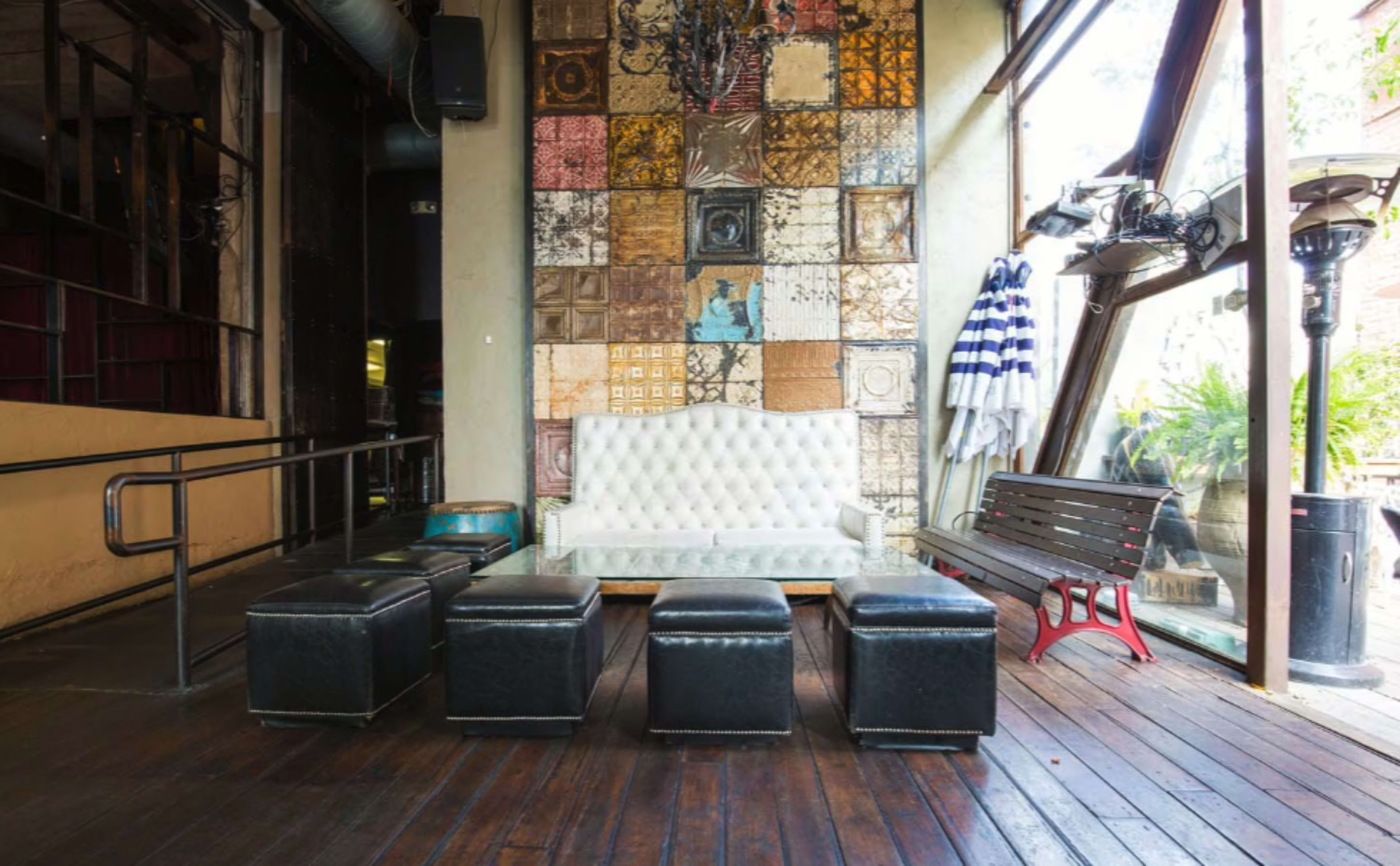 The scene features a cozy lounge area with a white tufted sofa, a glass coffee table, and four black ottomans arranged on a wooden floor, accompanied by a decorative wall of colorful metal panels.